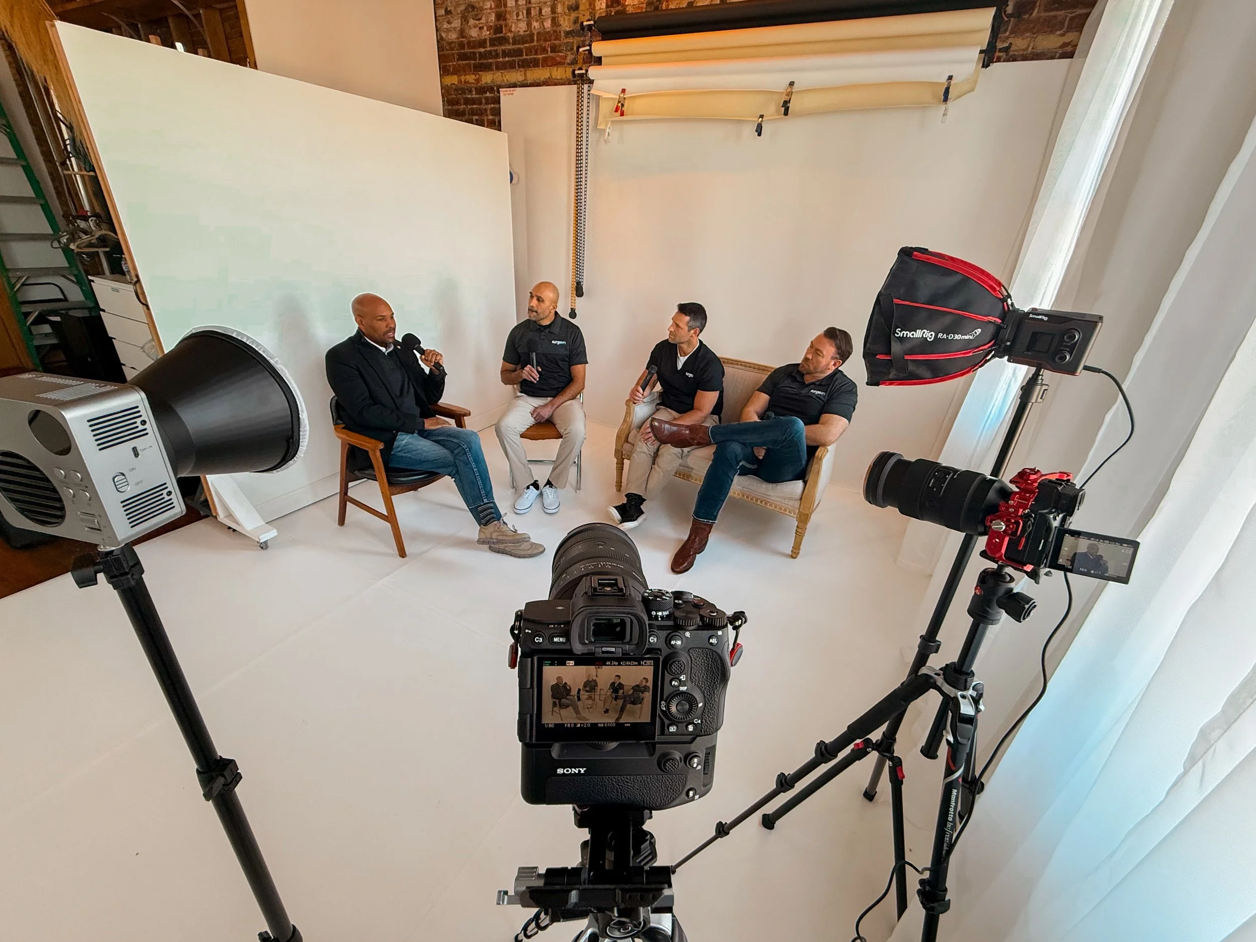 A group of four men sitting in a recording studio during an interview or discussion, with cameras and lighting equipment set up around them.