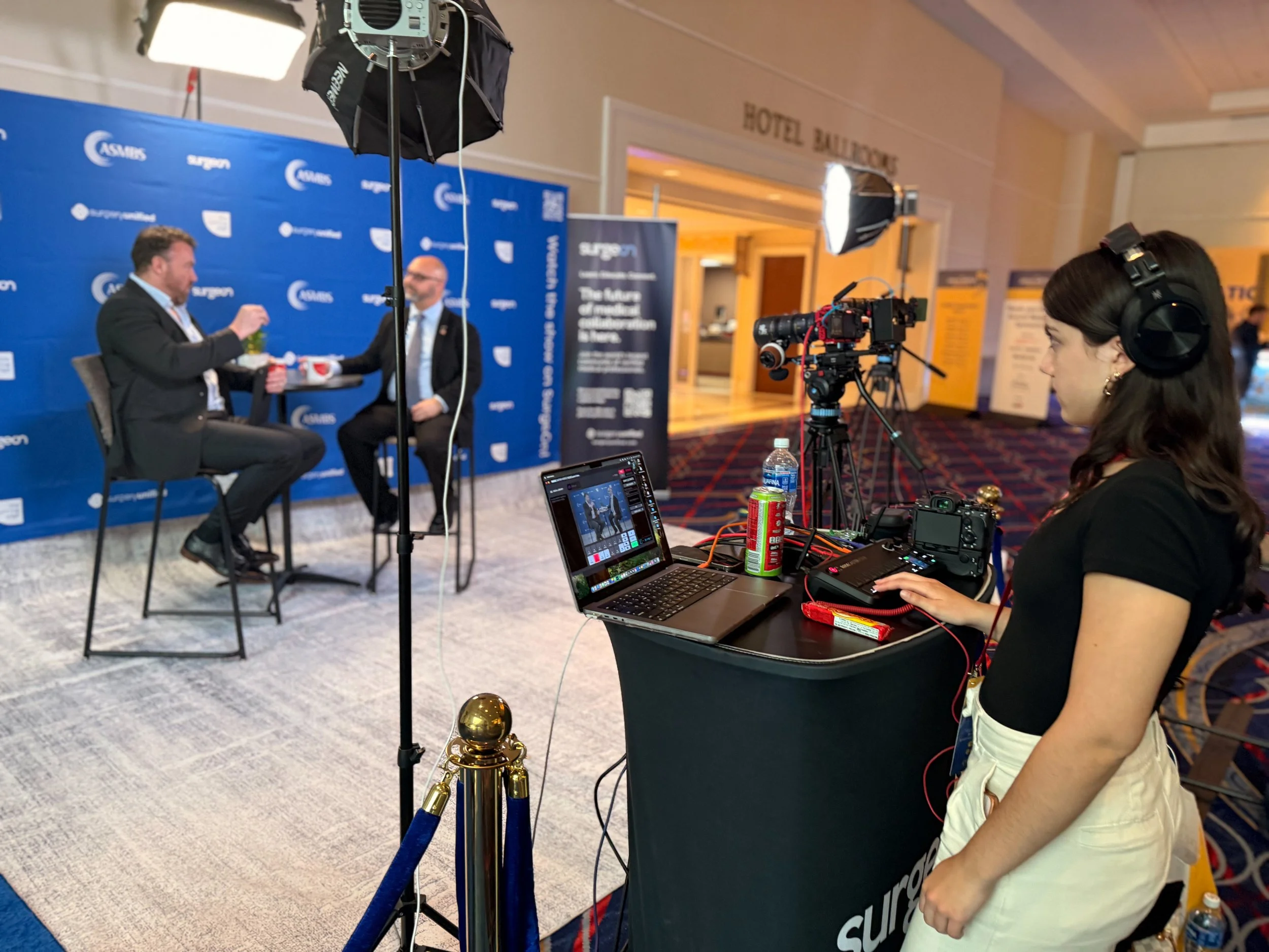 A woman operating a camera and laptop at a video recording setup with two men in suits being interviewed on stage in front of a blue backdrop with logos, inside a hotel conference hall.