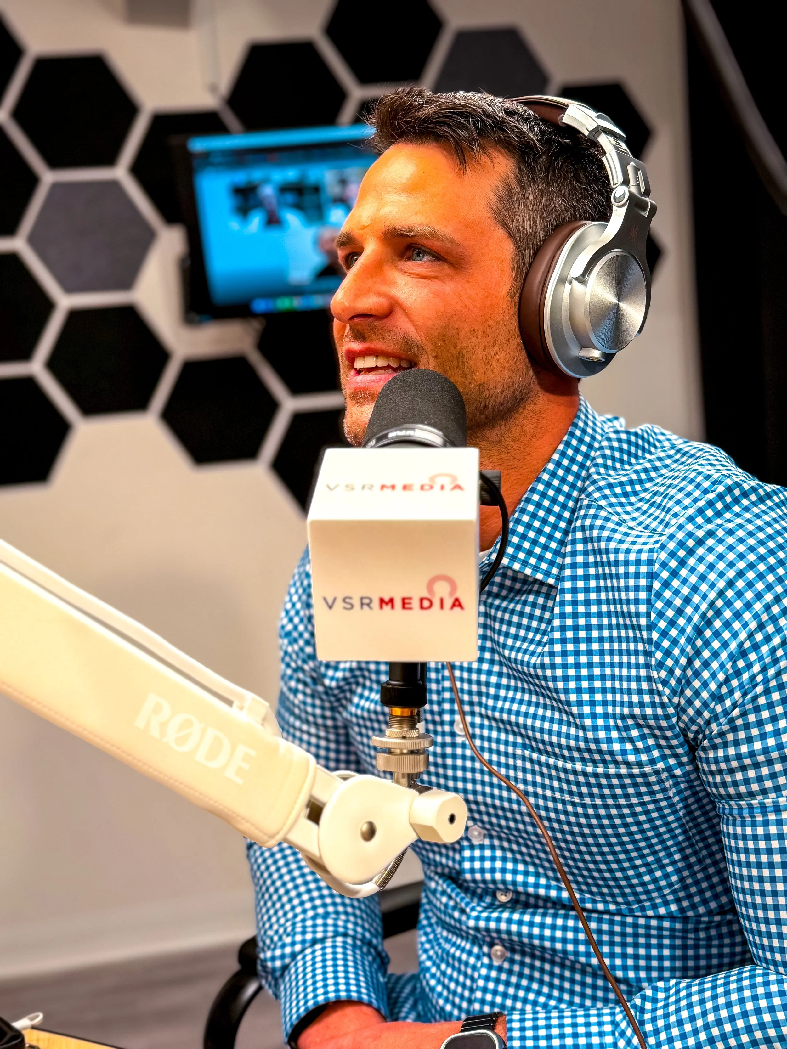 A man with short dark hair wearing headphones and a blue checkered shirt speaking into a microphone in a recording studio with a black and white hexagonal patterned wall and a monitor in the background.