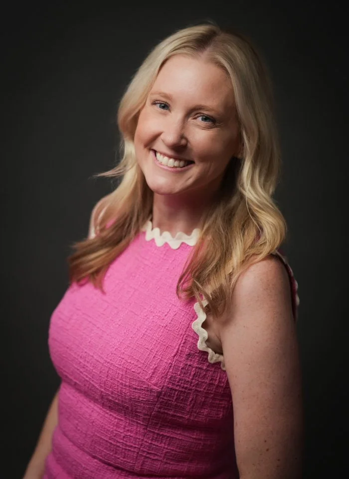 A woman standing in front of an abstract red and pink floral art background, smiling with one hand on her hip.