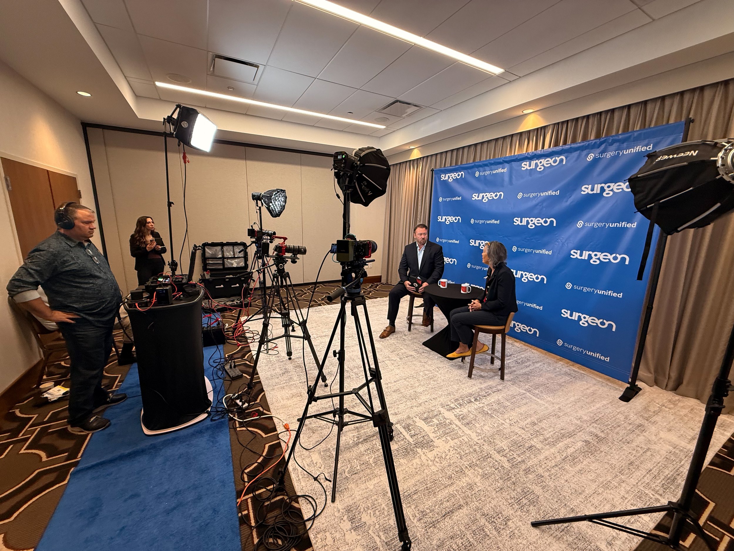 A behind-the-scenes view of a video interview setup in a conference room. Two individuals are seated at a table with microphones and coffee mugs, engaging in conversation against a blue "surgery" backdrop. Camera crew and lighting equipment are prese