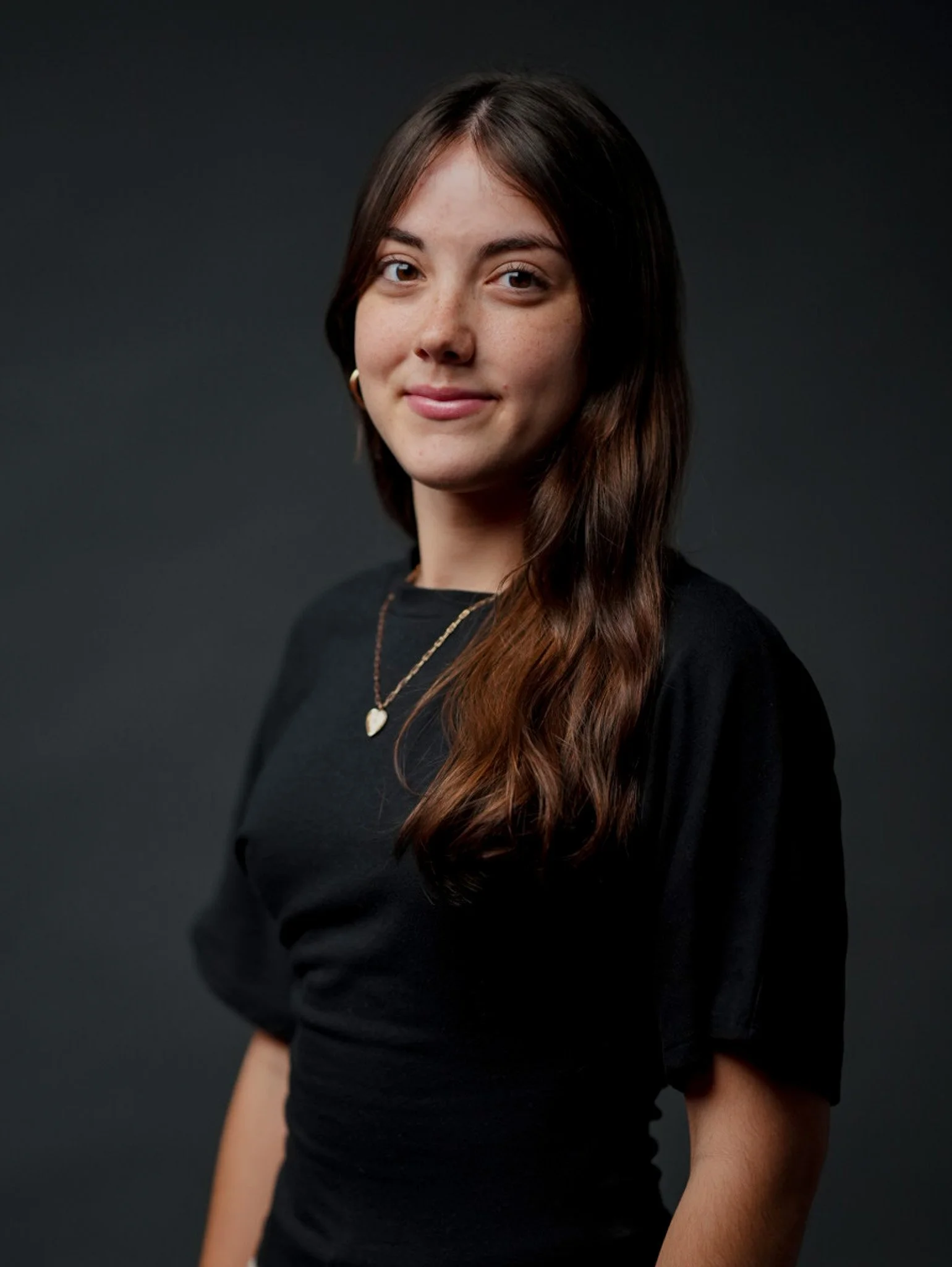 A young woman with shoulder-length wavy brown hair, smiling at the camera, wearing a black sleeveless top with white polka dots and a gold necklace, standing against a plain light gray background.