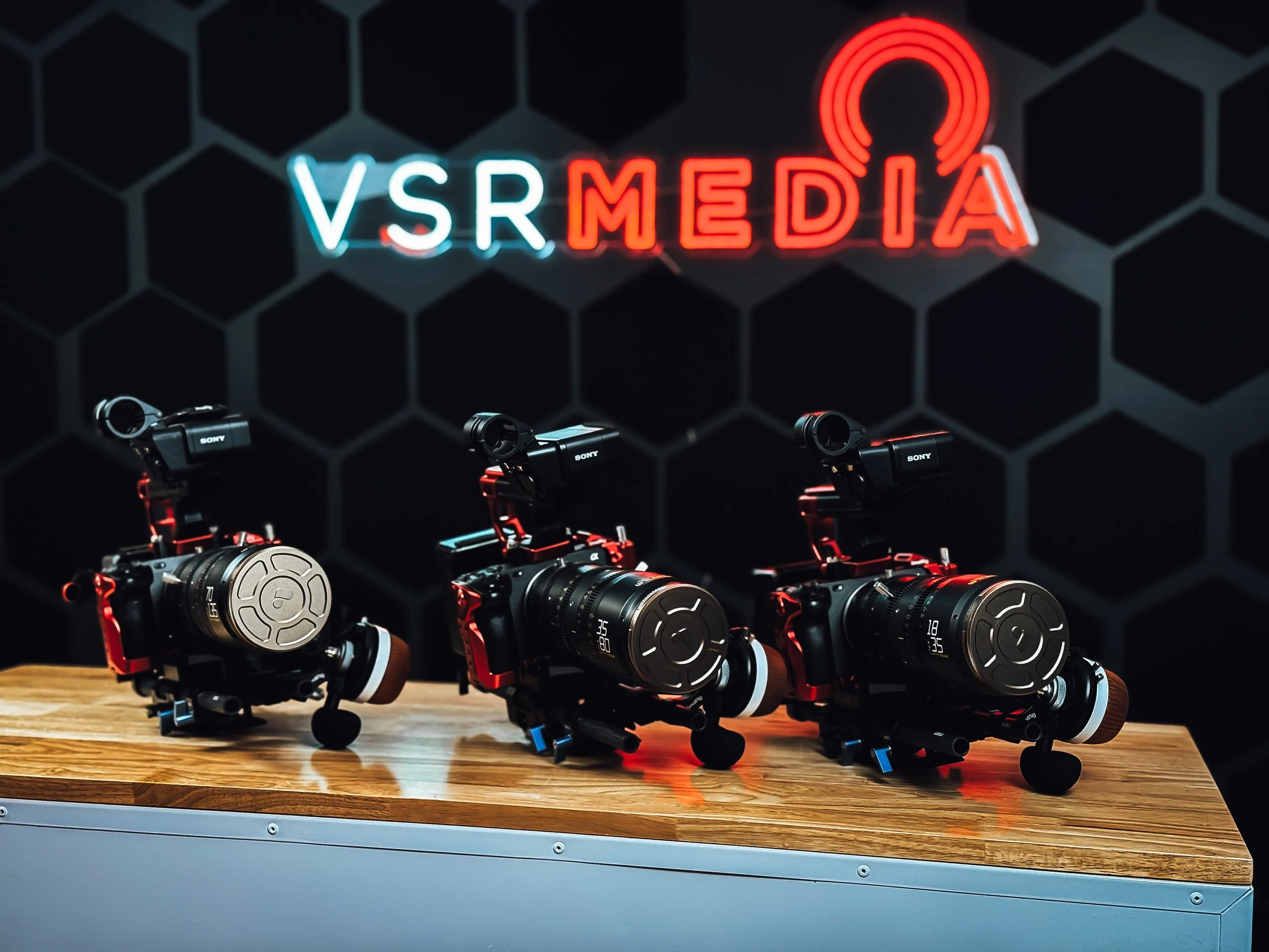 Three professional video cameras placed on a wooden table in front of a black honeycomb patterned wall with a neon sign that reads 'VSR MEDIA' in blue and red lights.