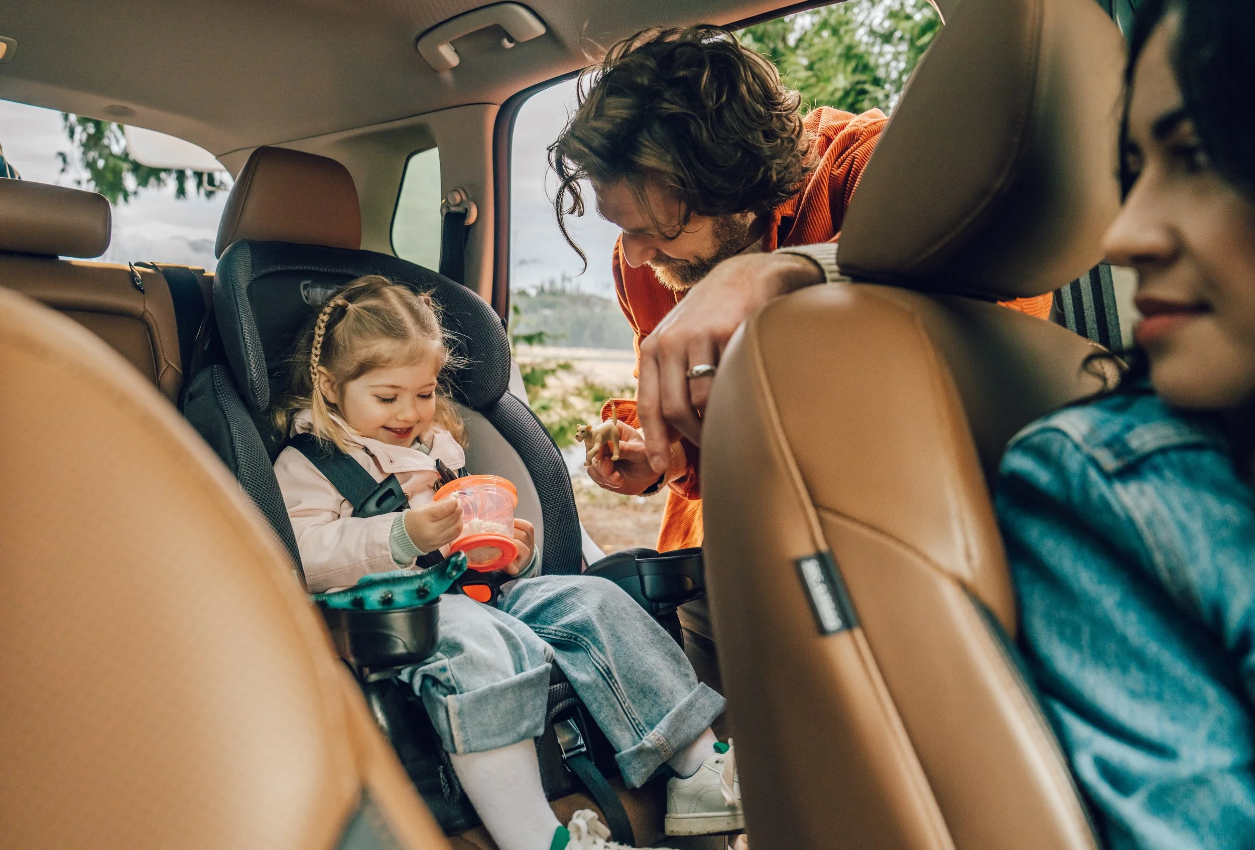 A man is leaning into a child safety car seat in the back of a vehicle, interacting with a young girl who is holding a toy and a cup.
