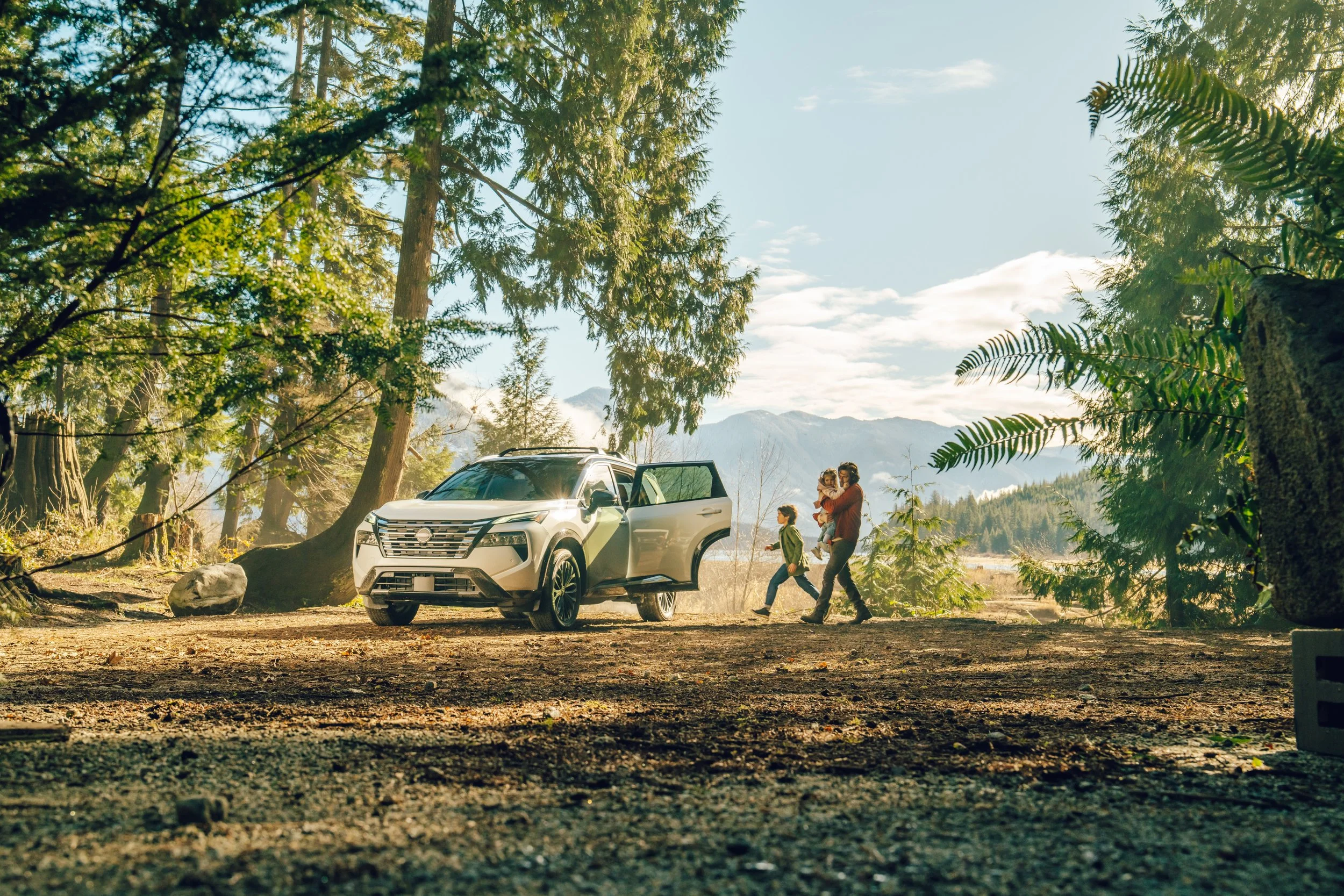 A family enjoying a walk in a forested area near a white SUV with its door open, with mountains visible in the background and sunlight filtering through the trees.