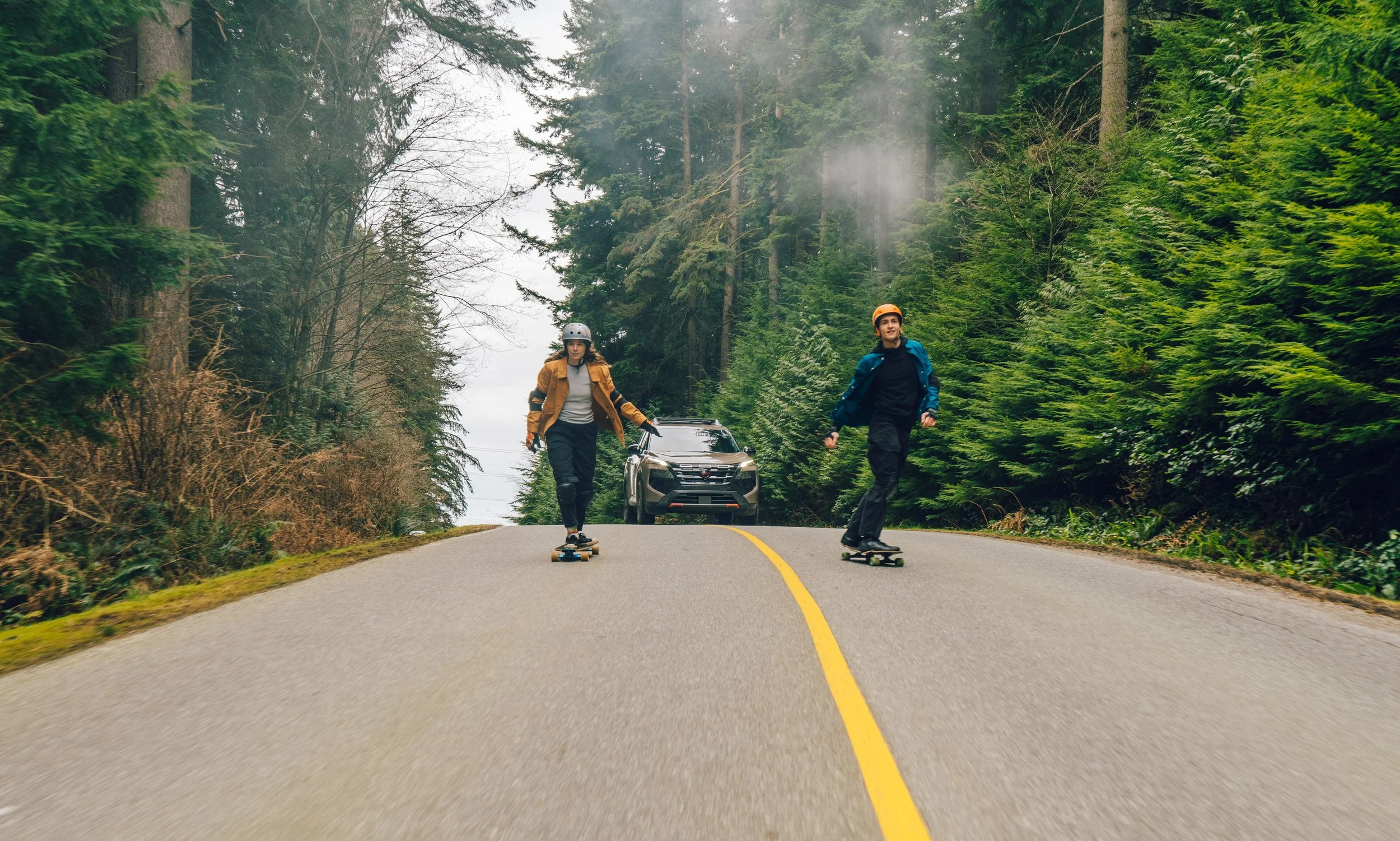 Two young people skateboarding on a winding forest road with tall pine trees surrounding them, and a parked car visible in the background.