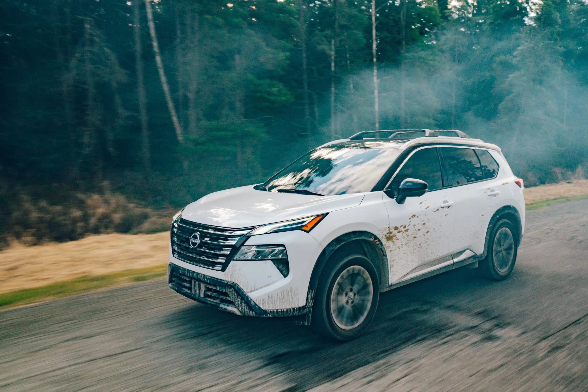 A white Nissan SUV driving on a road through a forest, kicking up dirt, with trees in the background and a slight motion blur.