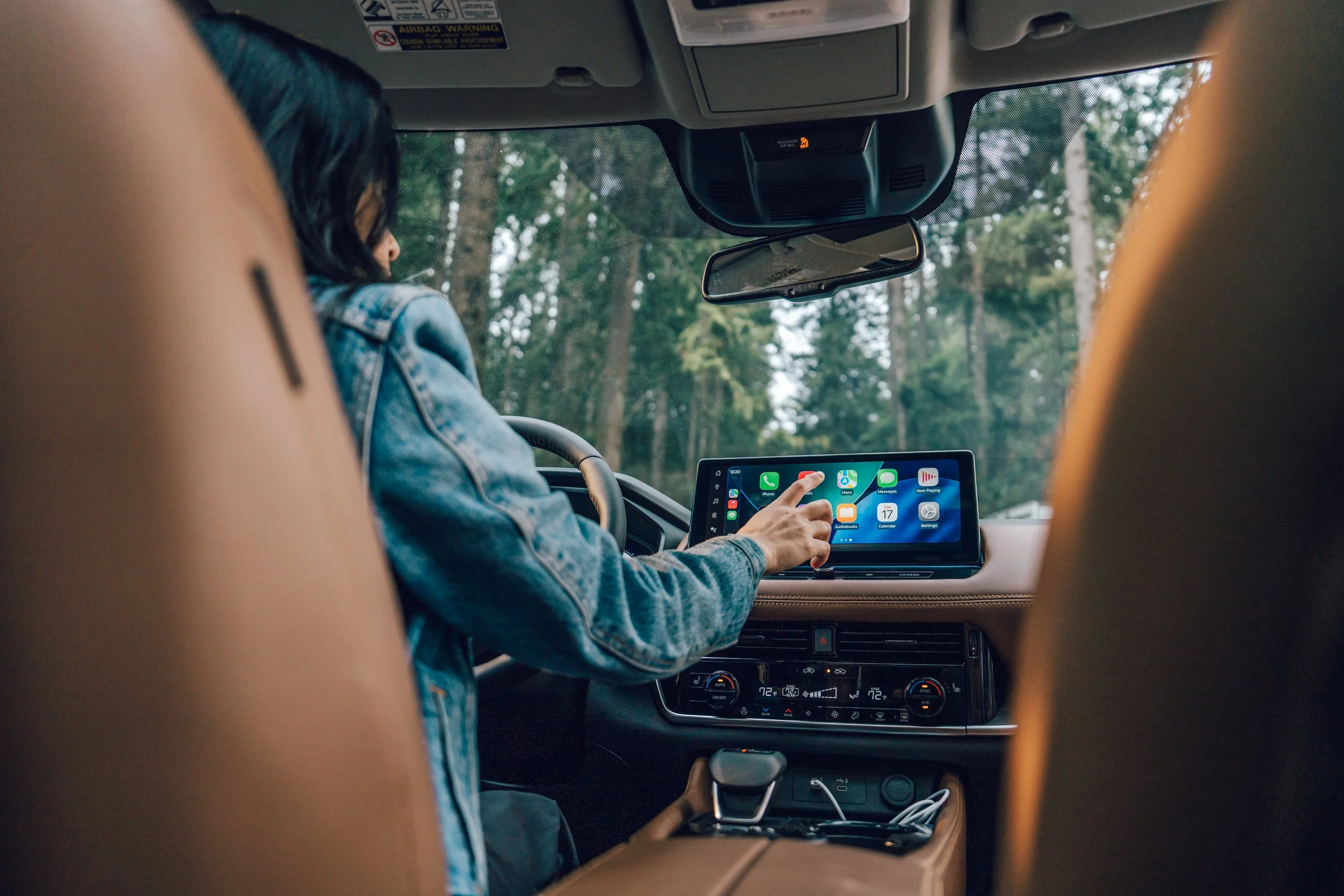 Inside a car, a woman with dark hair is sitting in the passenger seat, wearing a denim jacket and using a tablet device with colorful app icons, while the driver is partially visible in the foreground. The car's dashboard and rearview mirror are also