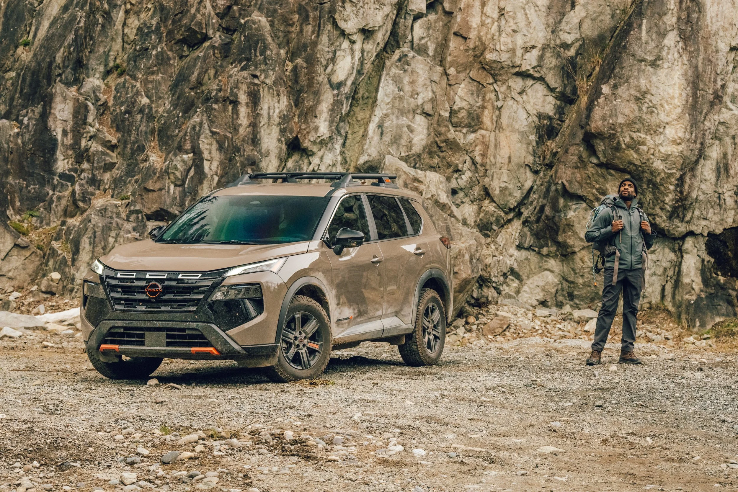 A man with a backpack standing next to a tan Nissan SUV parked on rocky terrain in front of a large rock wall, during a hiking or outdoor adventure.