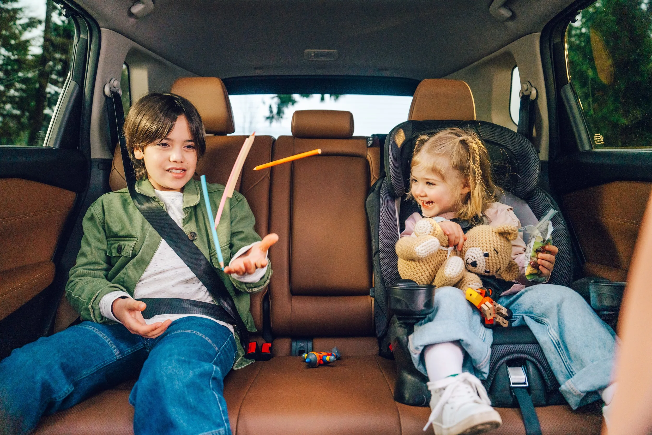 Two children laughing and talking inside a car's backseat, one boy and one girl, with toys and snacks around them.