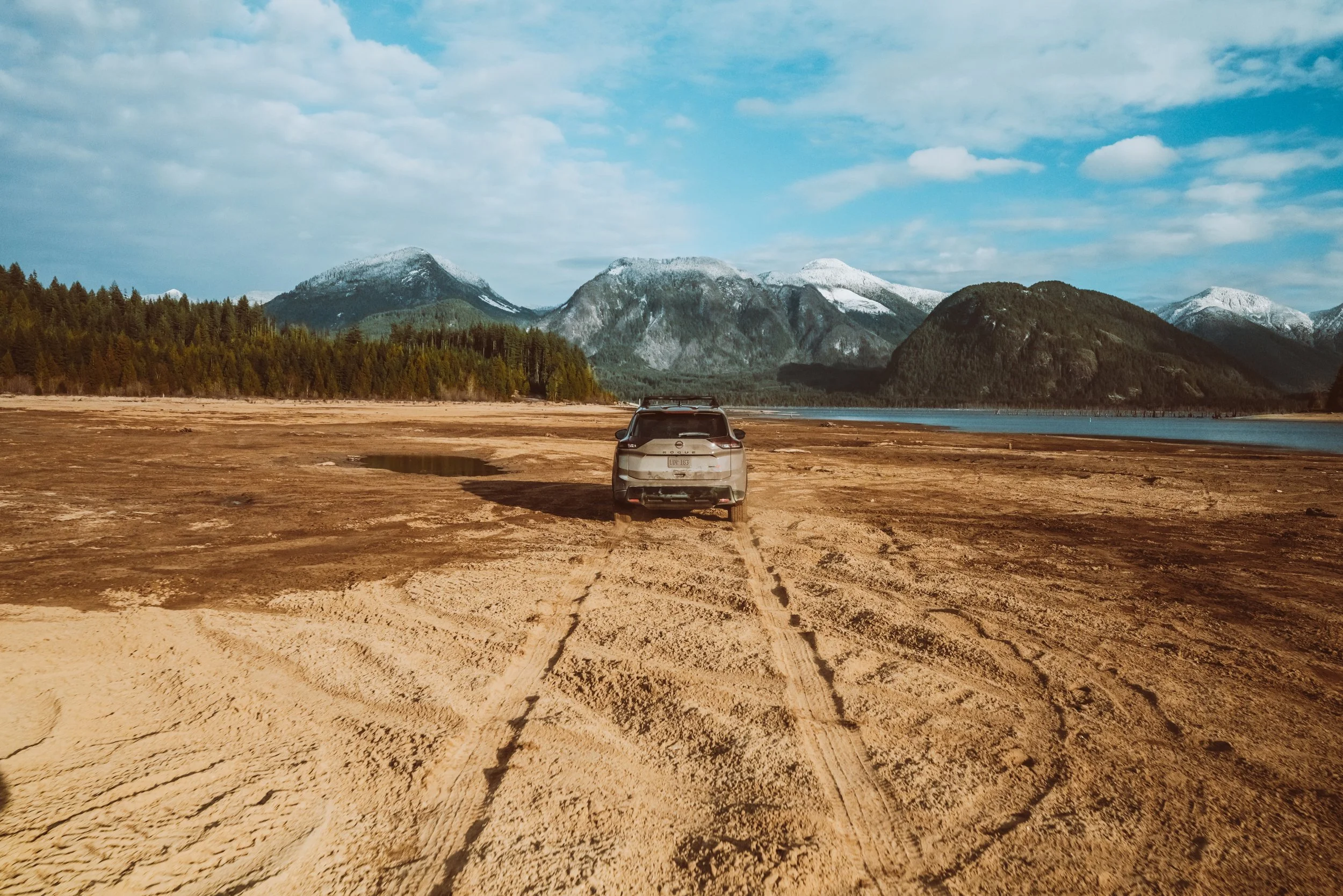 A car parked on a dirt clearing by a lake, with mountains in the background and a cloudy sky overhead.