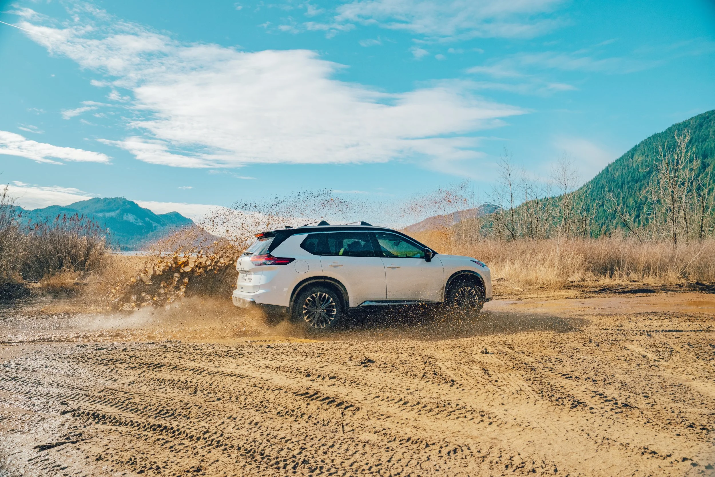 A white SUV driving through a muddy dirt road in a mountainous area, kicking up mud and water with a backdrop of green mountains and a partly cloudy blue sky.