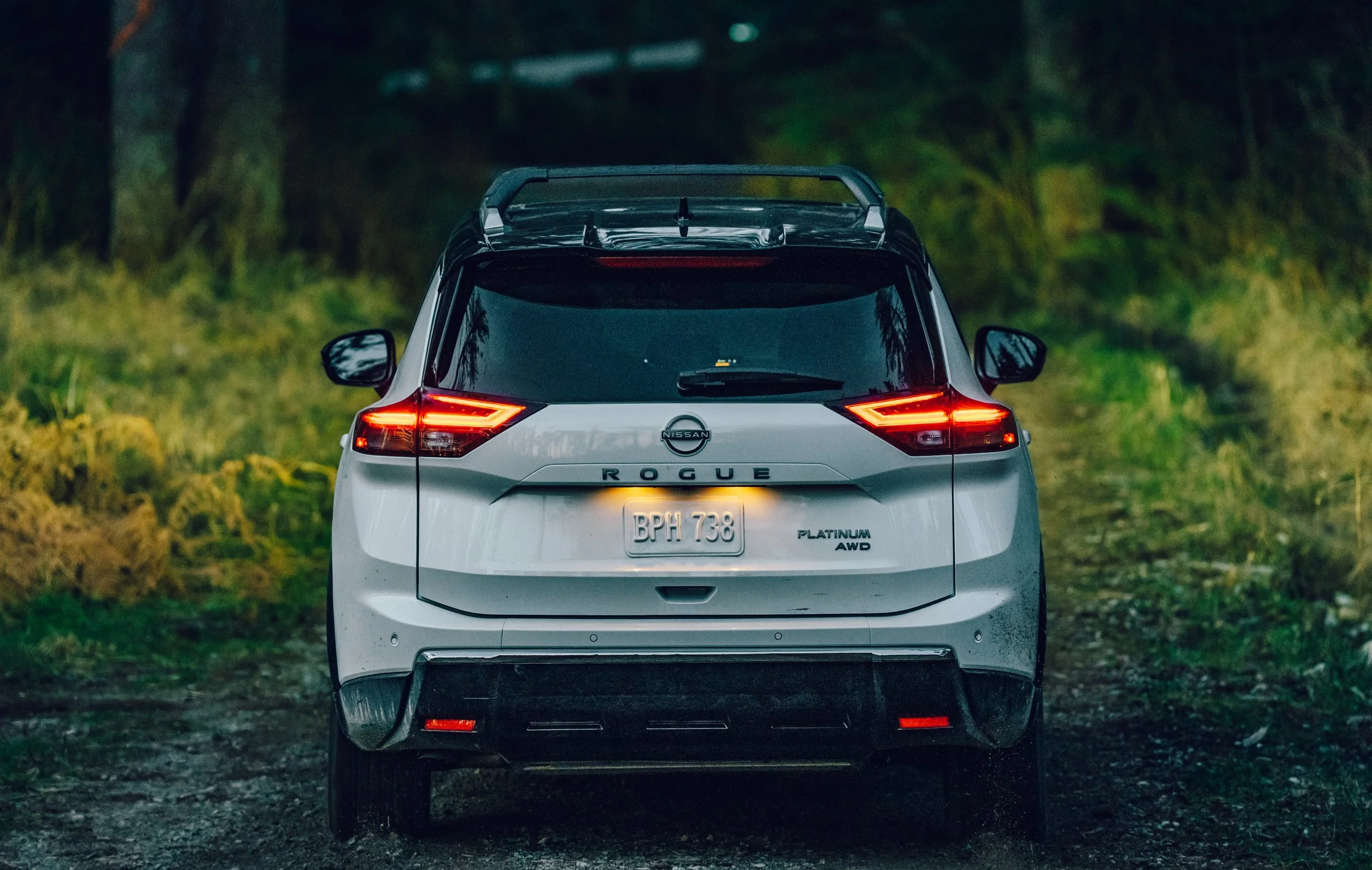 Rear view of a white Nissan Rogue SUV parked on a dirt or gravel path surrounded by green trees and foliage during dusk or evening.