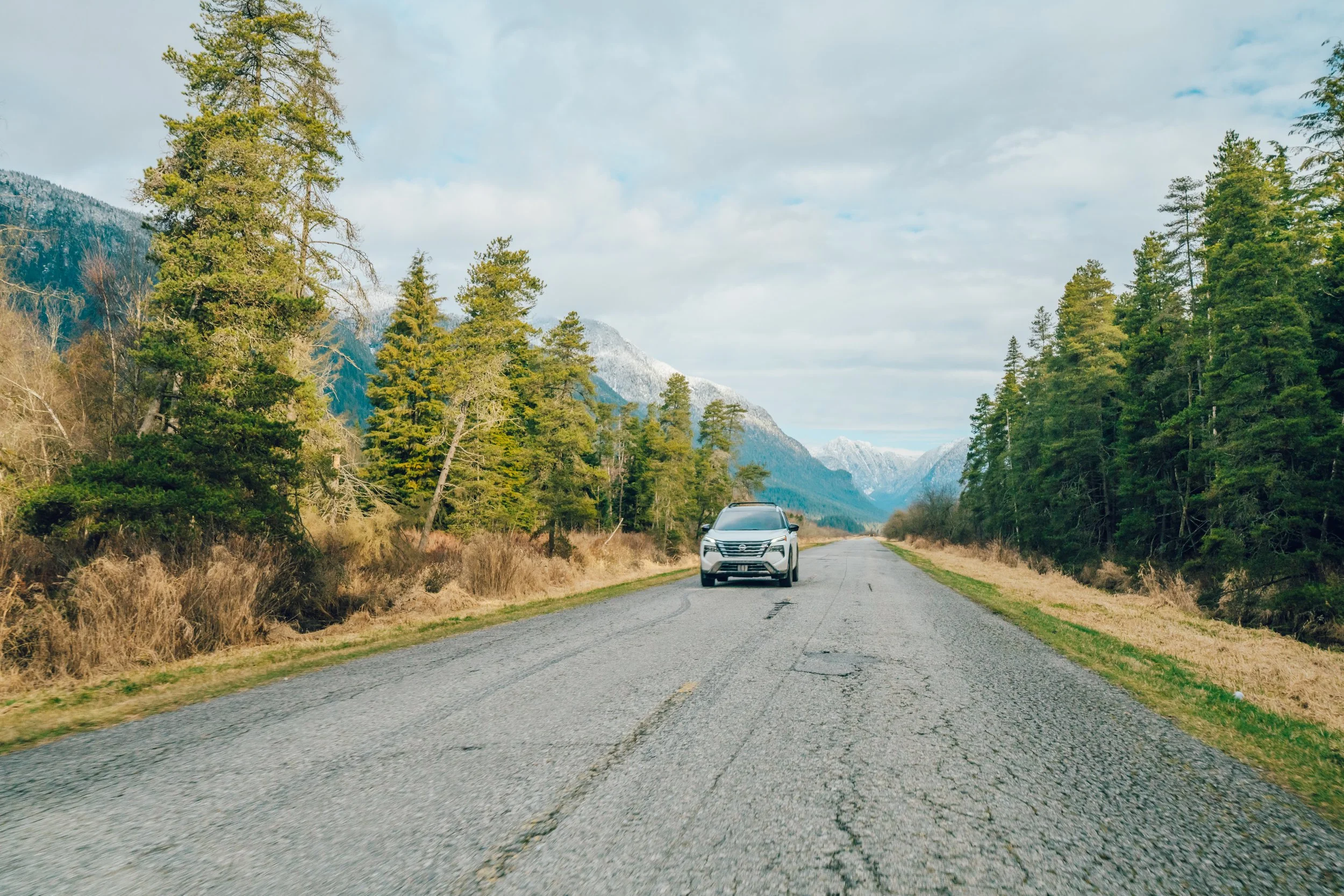 A silver SUV driving on a rural mountain road bordered by tall evergreen trees, with snow-capped mountains and a cloudy sky in the background.