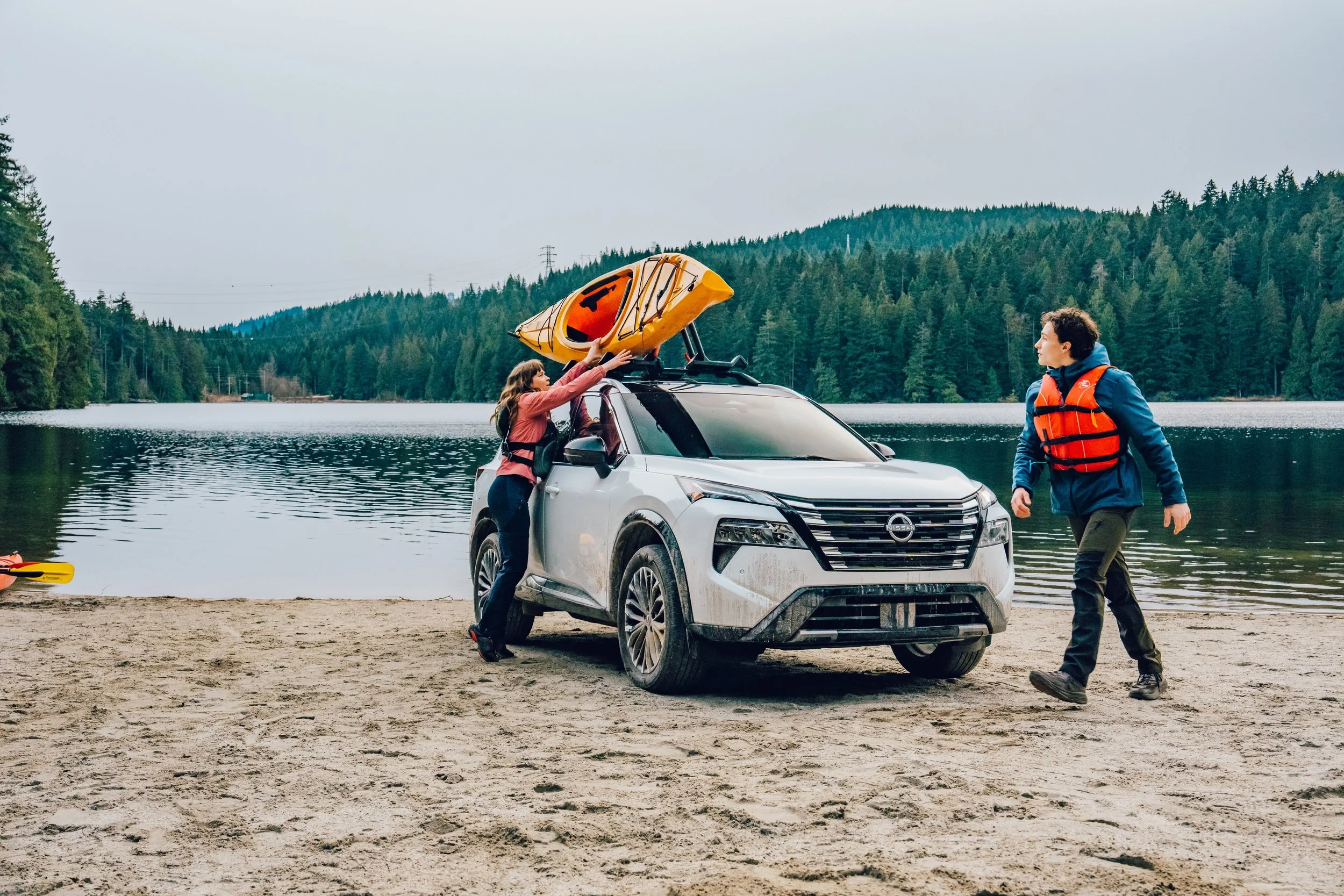 Two people preparing a kayak on top of a white Nissan SUV parked by a lakeside. One person is handling the kayak while the other is walking nearby. The scene includes a sandy shore, a large body of water, and a forested hillside under an overcast sky