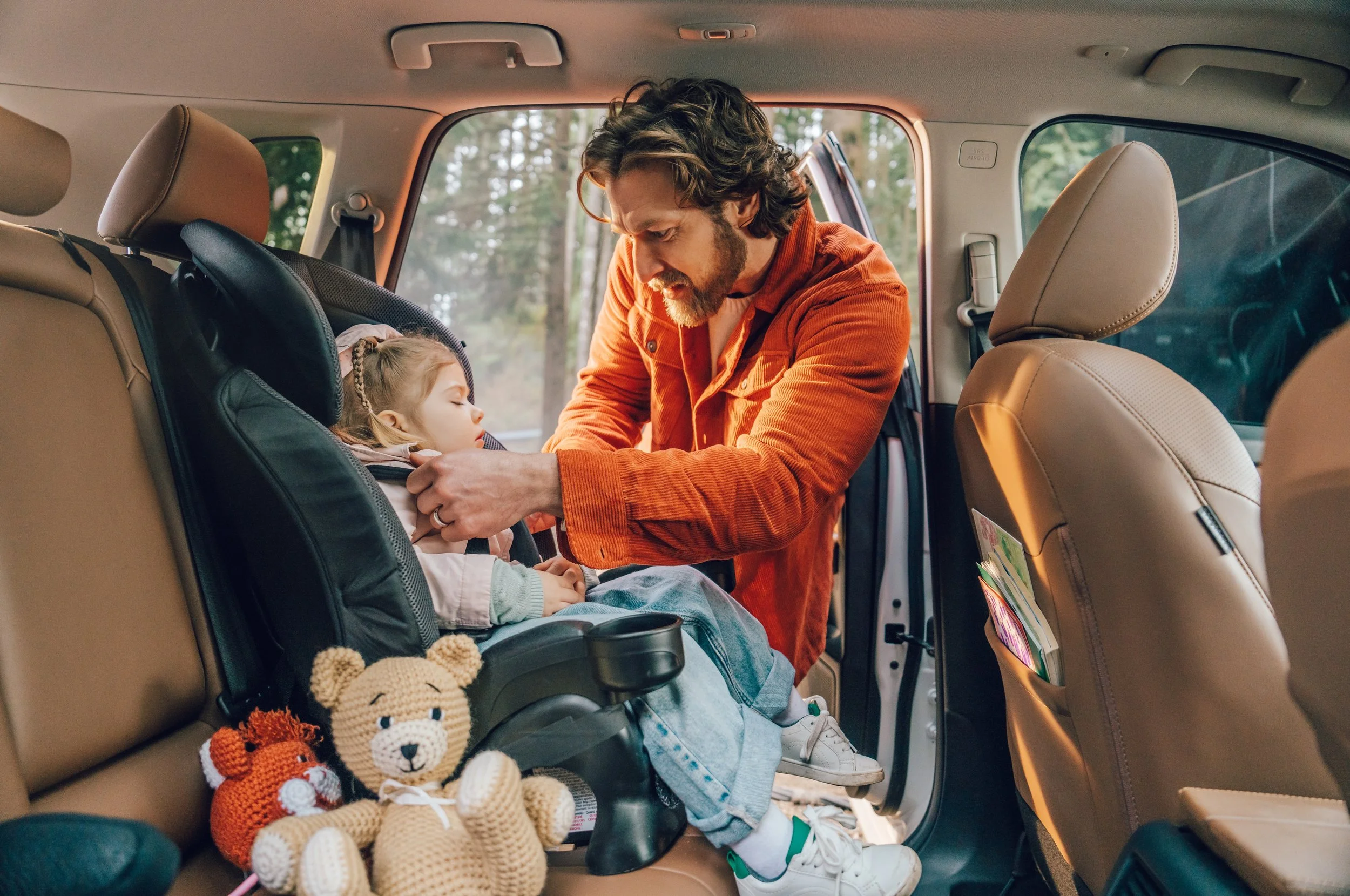 A man with curly hair and a beard adjusting a young girl with a braids in a car seat inside a vehicle, with toys including a crocheted lion and fox on the seat.