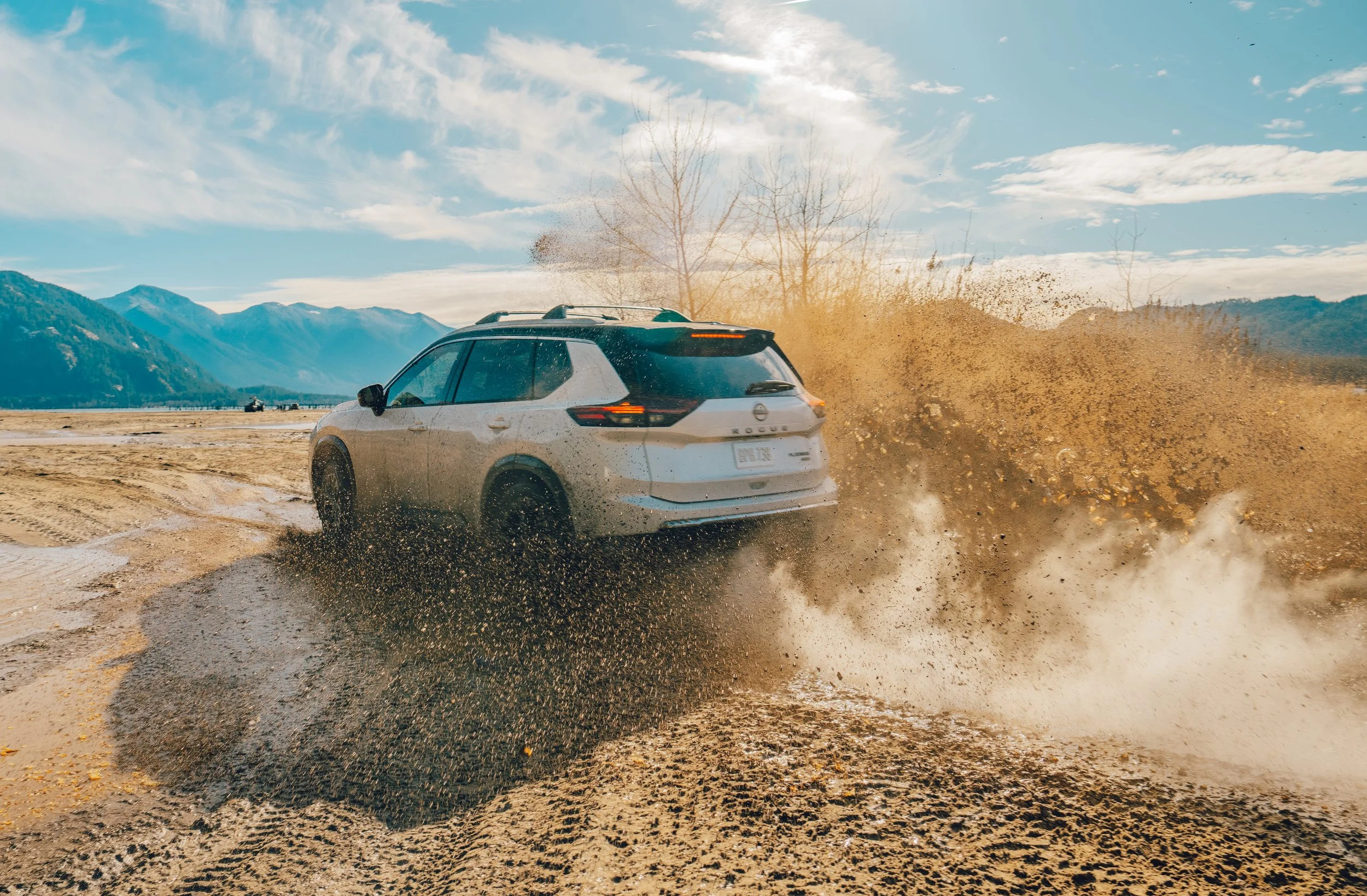 A white SUV driving fast through a muddy puddle, splashing water and dirt into the air, with mountains and a partly cloudy sky in the background.