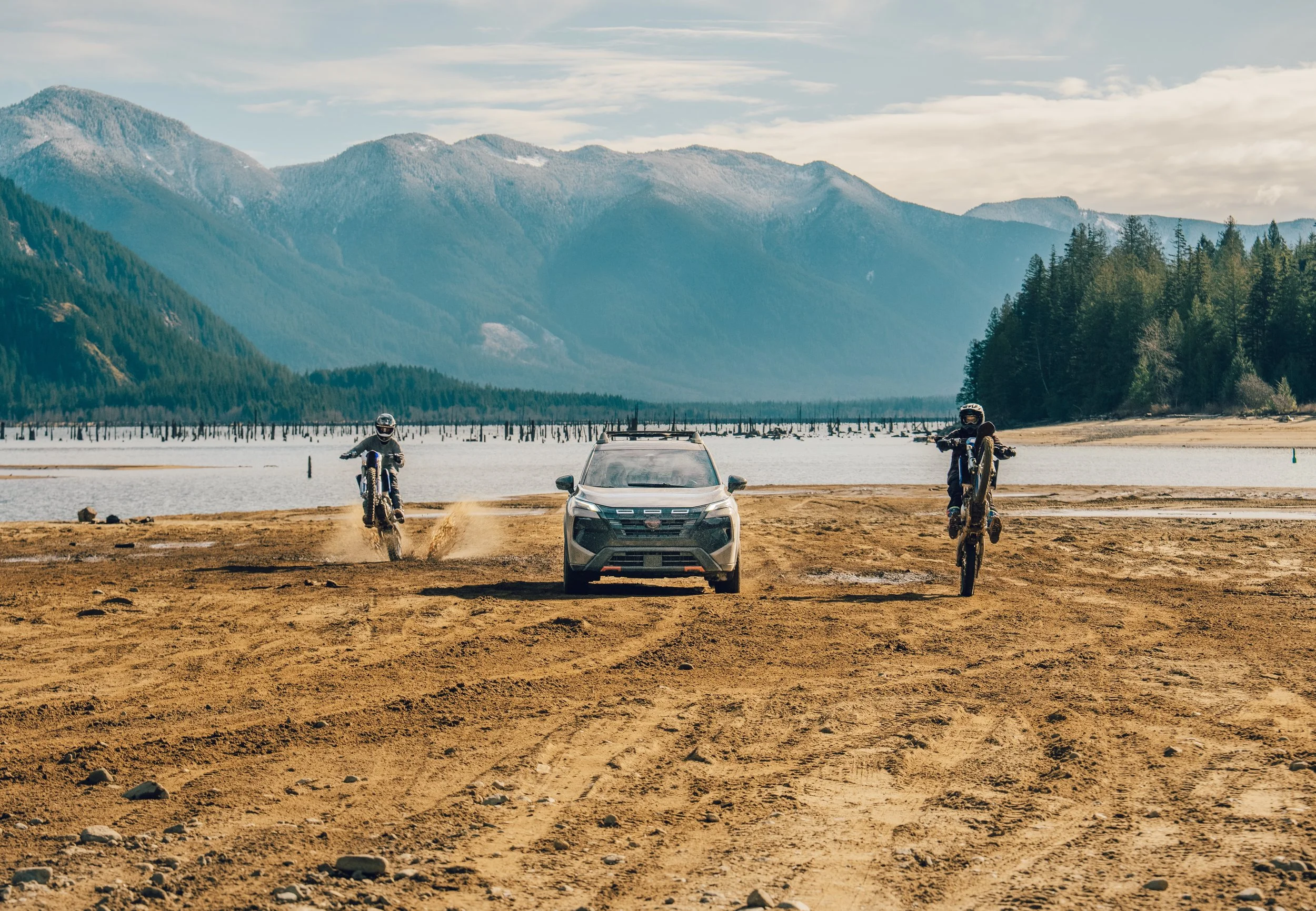 Two motorcyclists performing wheelies on dirt bikes on a sandy, open area by a lake, with mountains in the background under a partly cloudy sky, and a silver car parked between them.