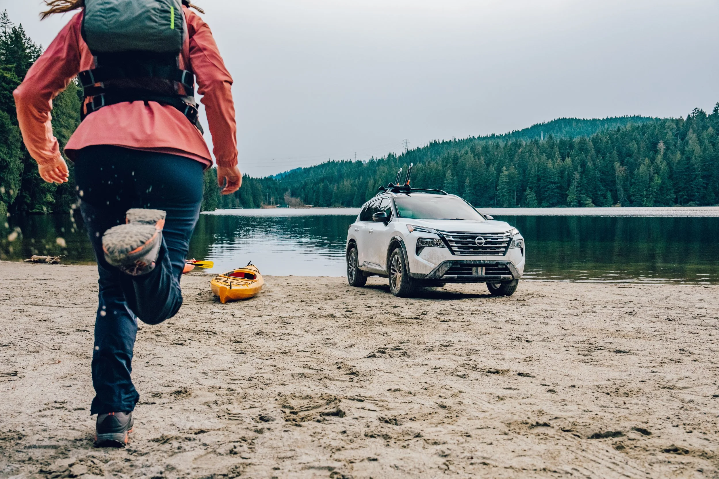 A person jogging on a sandy beach near a body of water with kayaks, a white SUV parked on the beach, and a forested hillside in the background.