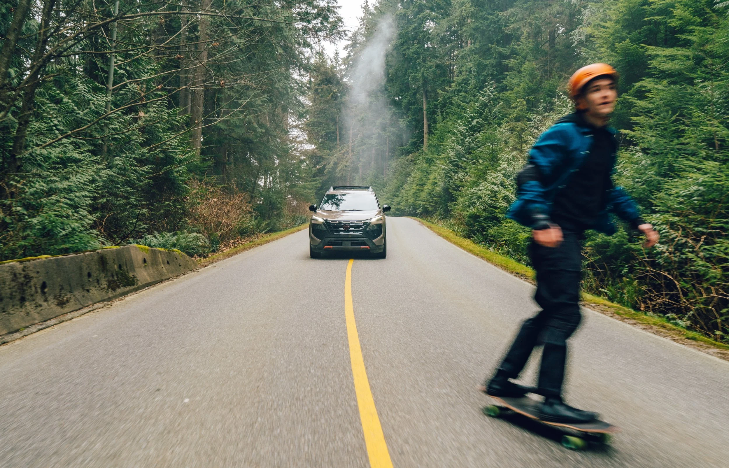 A person wearing a blue jacket and an orange helmet skateboarding on a narrow forest road with a car stopped in the background, surrounded by tall trees and greenery.