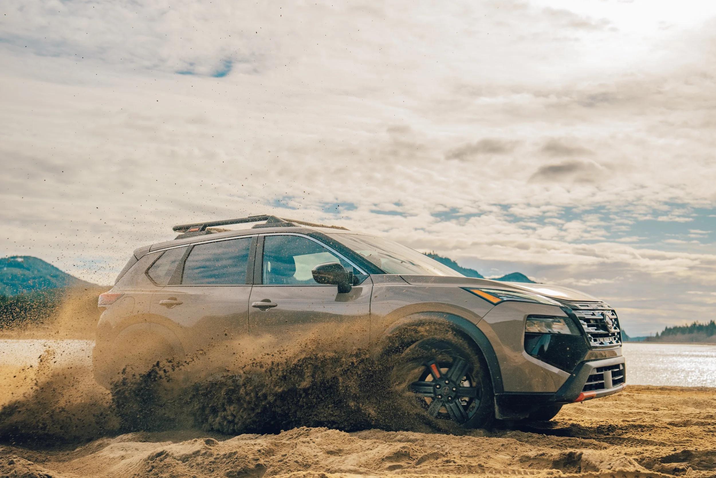 A silver SUV driving through a muddy terrain on a cloudy day with mountains and cloudy sky in the background.