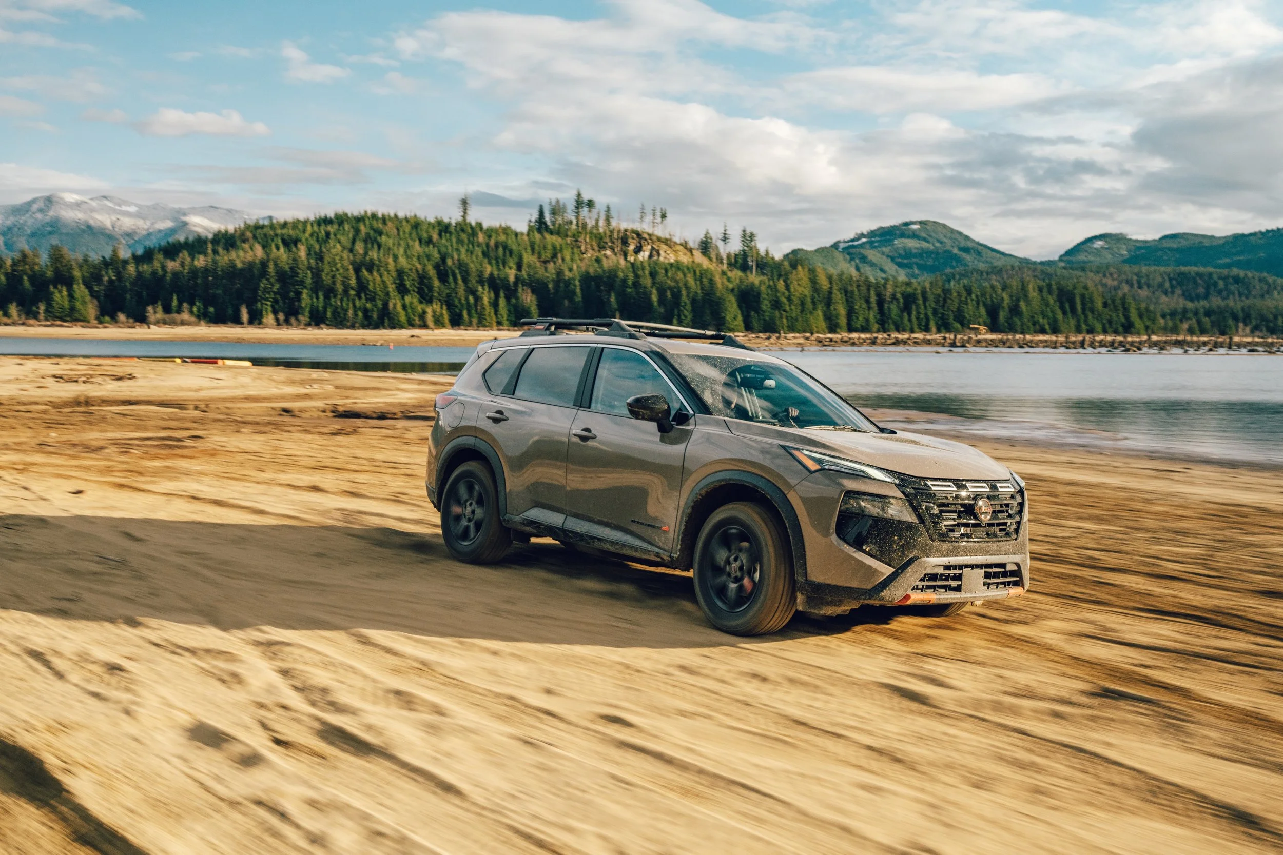 A gray SUV driving on a dirt road beside a body of water with forest and mountains in the background under a partly cloudy sky.