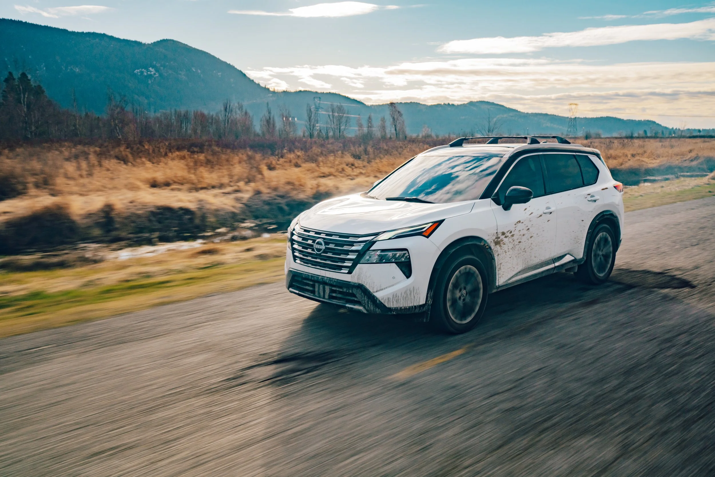 A white SUV driving on a rural road with mountains and a cloudy sky in the background.