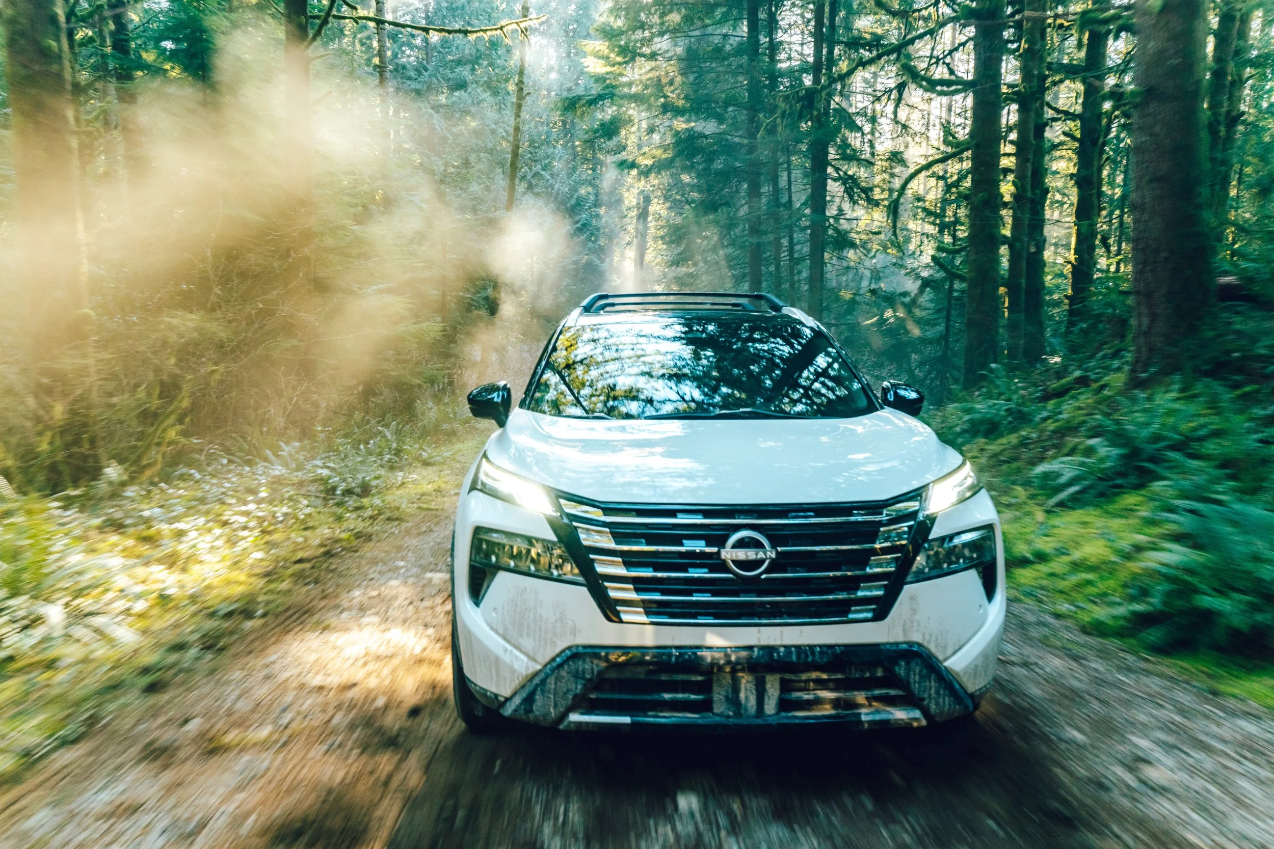 A white Nissan SUV driving through a forested dirt trail surrounded by tall green trees with sunlight filtering through.
