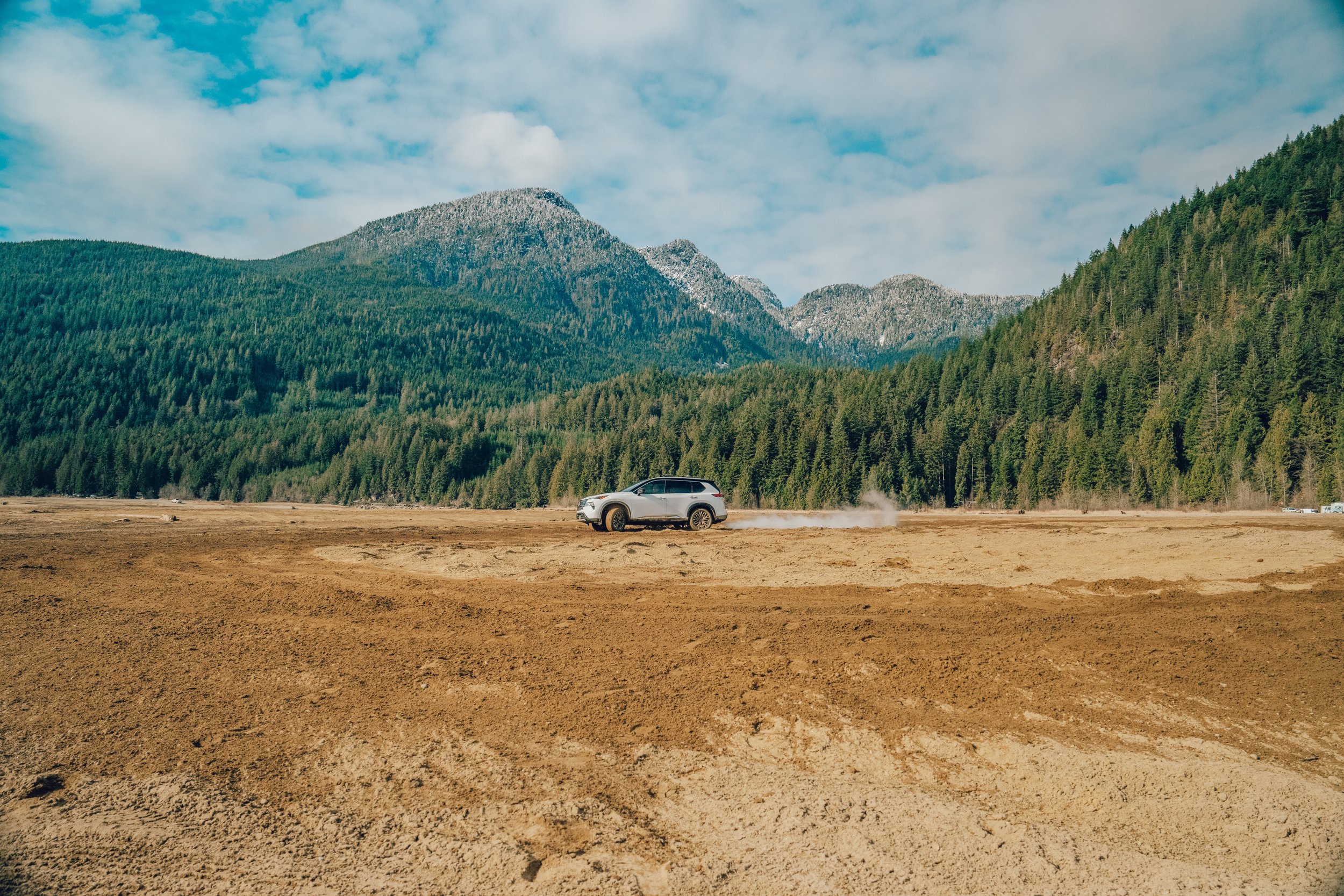 Silver SUV driving across a dry, dusty open field with dust clouds behind it, surrounded by dense green forests and mountain ranges under a partly cloudy sky.