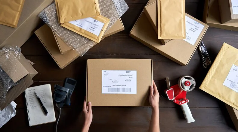 Hands packing a laptop shipping box with labels, bubble wrap, and packing supplies for a remote employee return