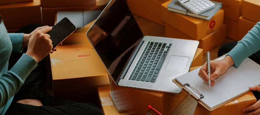 Two employees preparing laptop shipments with boxes, phone, and checklist — representing the remote offboarding guide and laptop retrieval process with shipping costs for domestic and international work-from-home equipment in 2026
