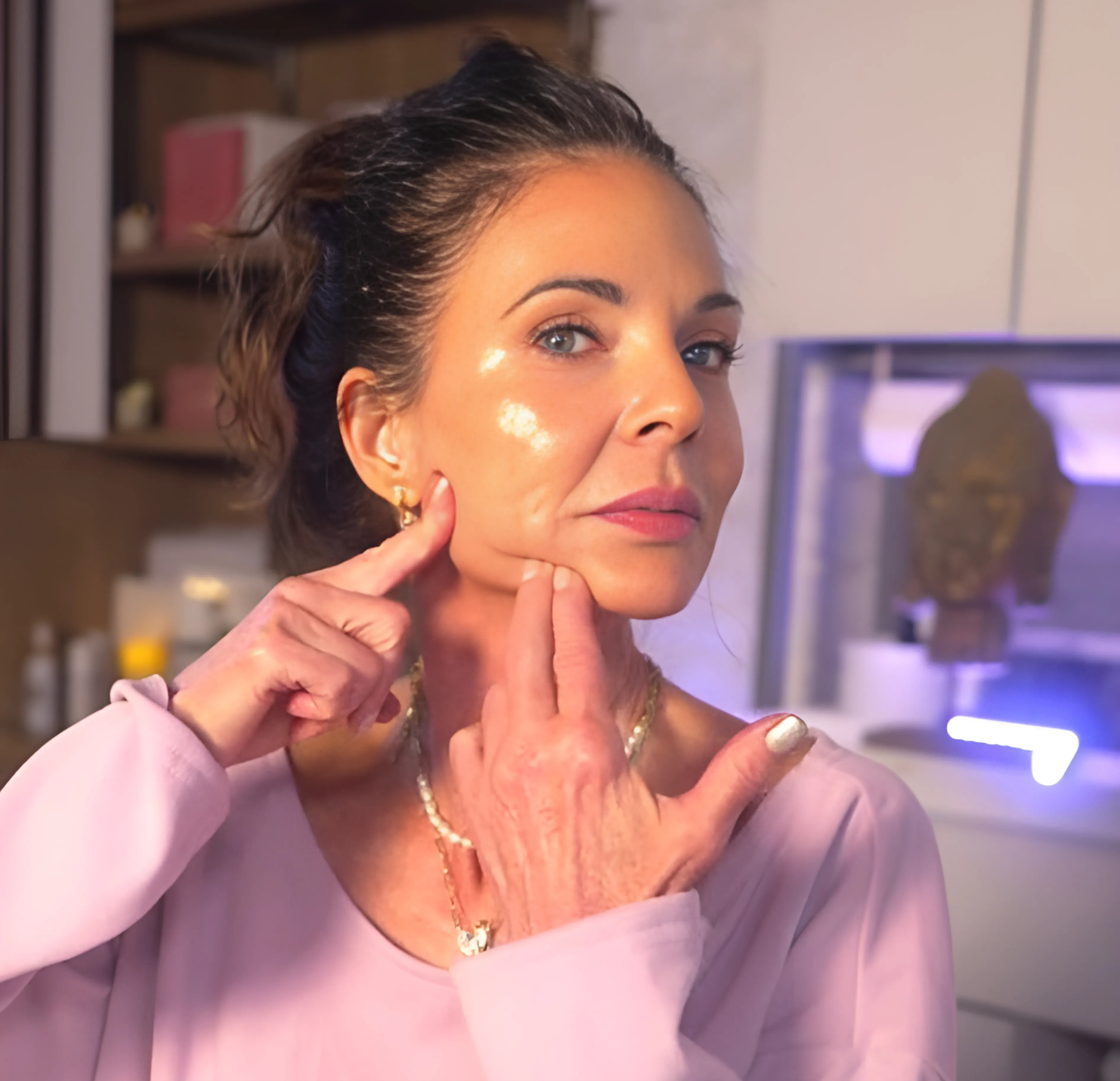 A woman with dark hair pulled back, touching her face near her cheek and chin, in a room with medical or aesthetic equipment in the background.
