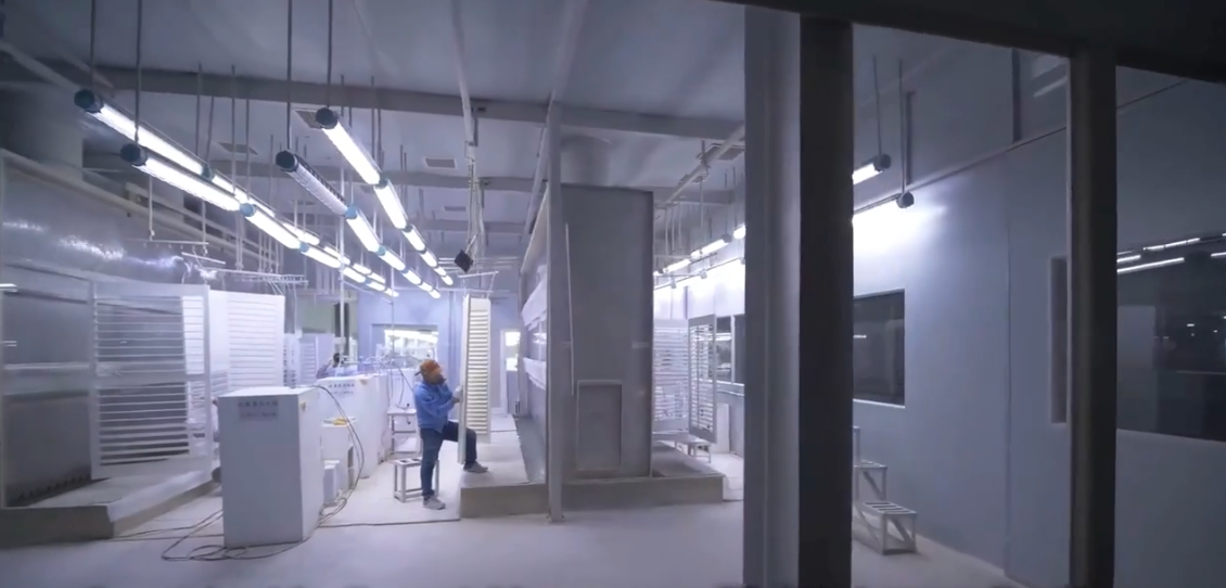 Interior of a clean, modern laboratory with white walls, fluorescent lighting, and scientific equipment. Two people are working with equipment on the left side.