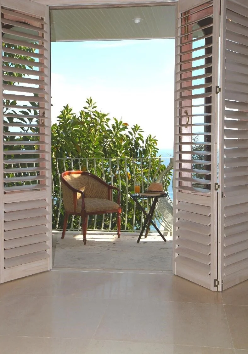 Balcony with a chair, a table with a drink and book, overlooking leafy trees and a distant view of water under a clear sky.