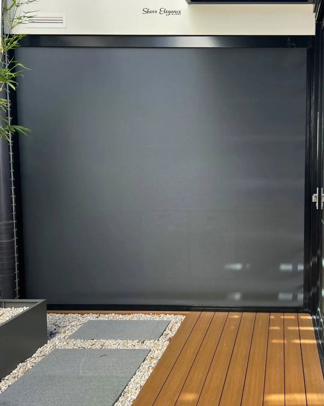 A modern outdoor space featuring a gray retractable screen or fence, small square gray outdoor mats on white pebble ground, a small potted plant, and a wooden deck in the corner. The top of the screen has the text 'Sheer Elegance'.