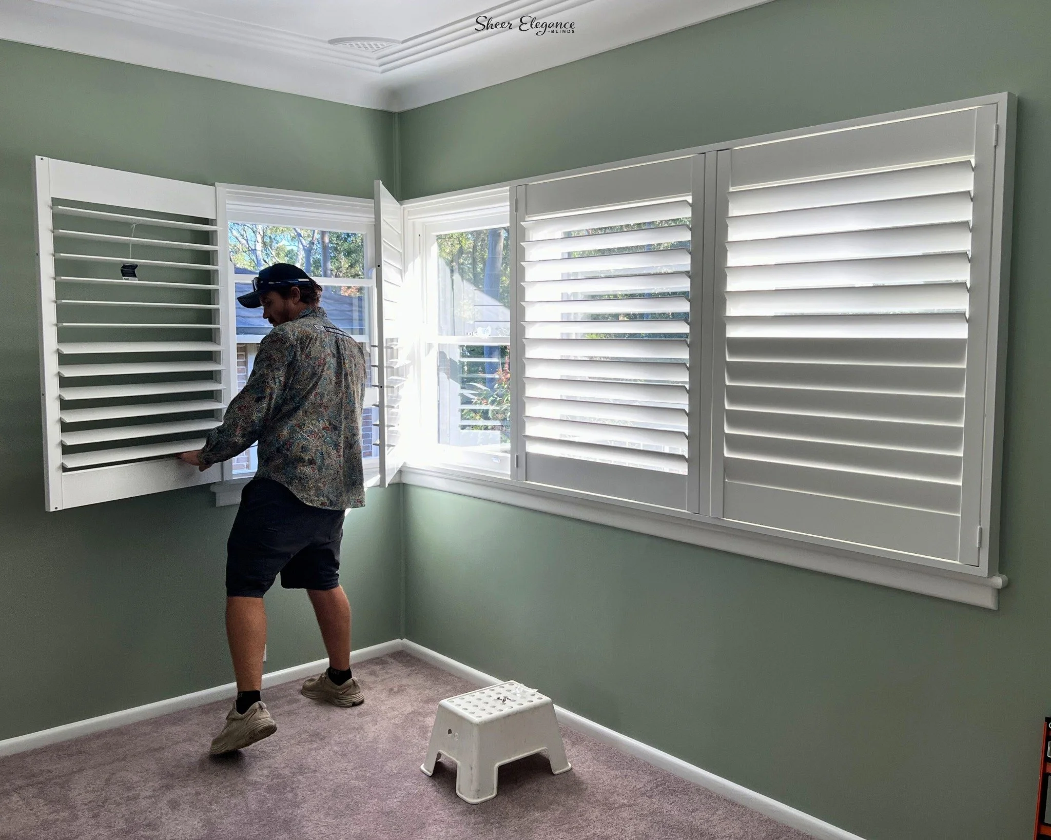 A person installing white plantation shutters on a window in a room with green walls and gray carpet. There is a small white step stool and portable heater in the room.