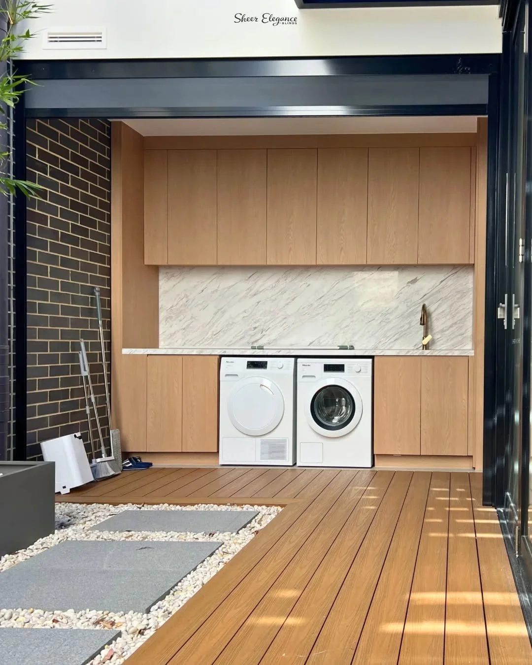 Modern laundry room with wooden cabinets, marble backsplash, washer, dryer, and gold faucet, on a wooden deck with stone pathway.
