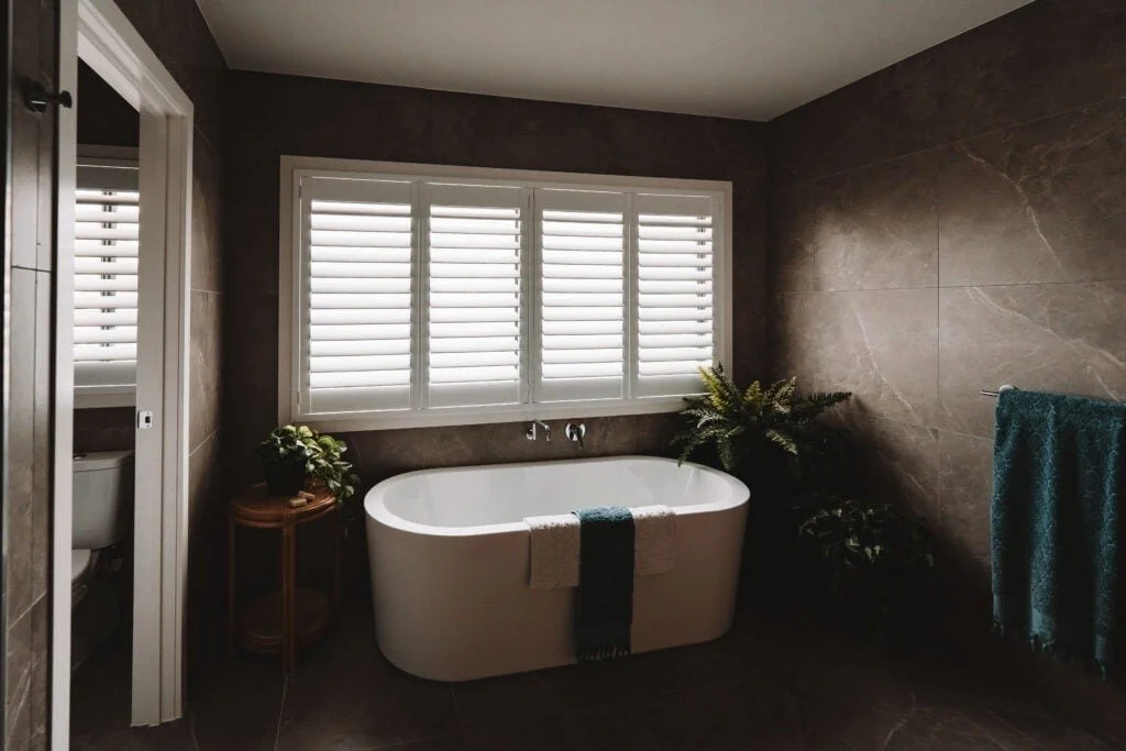 Bathroom with a free-standing bathtub, plant decorations, and large window with white shutters.