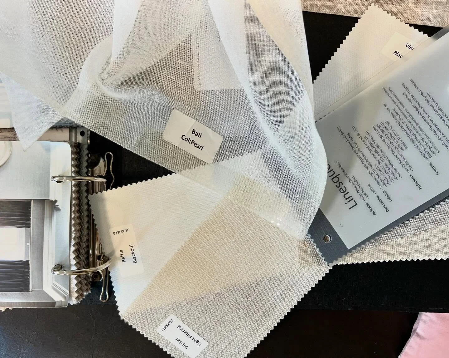 Fabric swatches labeled 'Bali Col: Pearl' and 'Light Beige' placed on a black surface. There are fabric sample cards, labels, and a clipboard with printed info. The swatches have pinking shears holes for hanging.