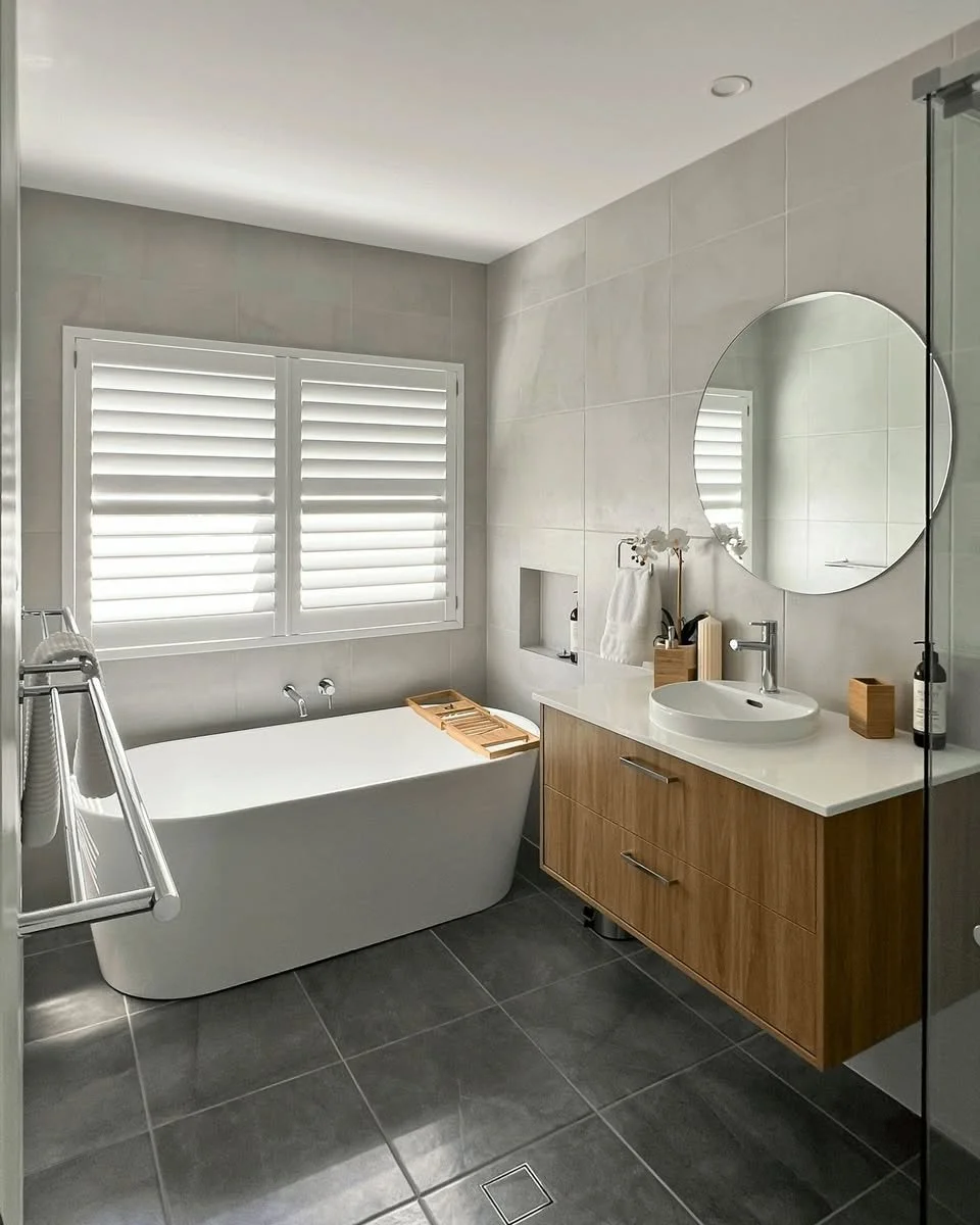 Modern bathroom with gray tiled floor and walls, a white bathtub, a window with white plantation shutters, a wooden vanity with a round mirror, and a white vessel sink.