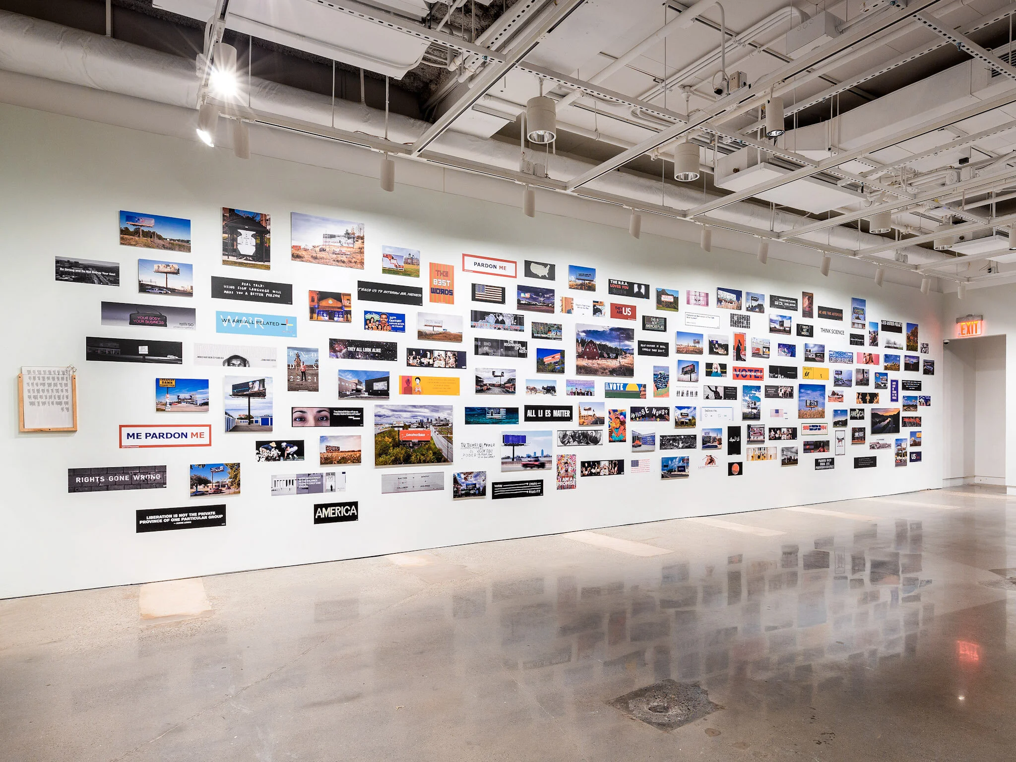 An art gallery wall displaying numerous small photographs, signs, and posters related to America and political messages, arranged in a grid pattern in a bright, modern space with a polished floor.