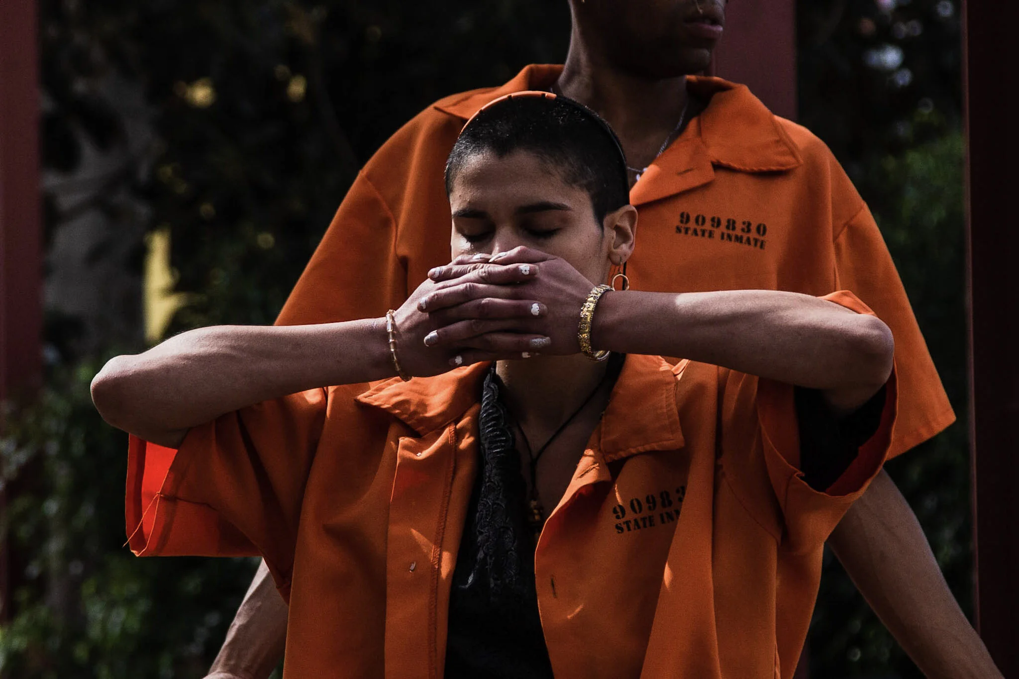 Person with short hair and earrings in orange prison jumpsuit with prisoner ID, covering nose and mouth with hands, standing outside with another person partially visible behind them.