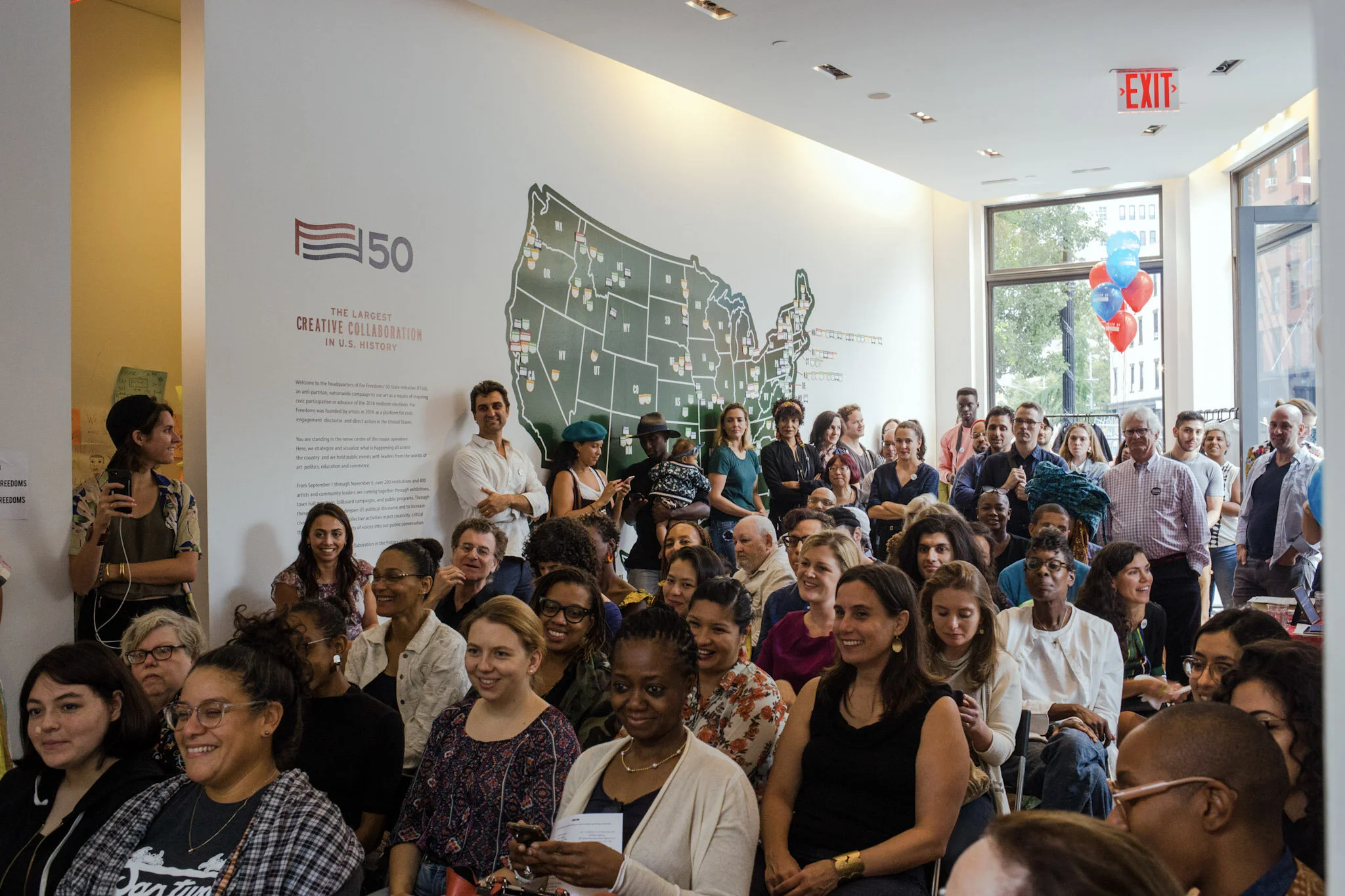 A diverse group of people attending a large event inside a room with a wall displaying a map of the United States and the number 50, celebrating U.S. history and civic participation.