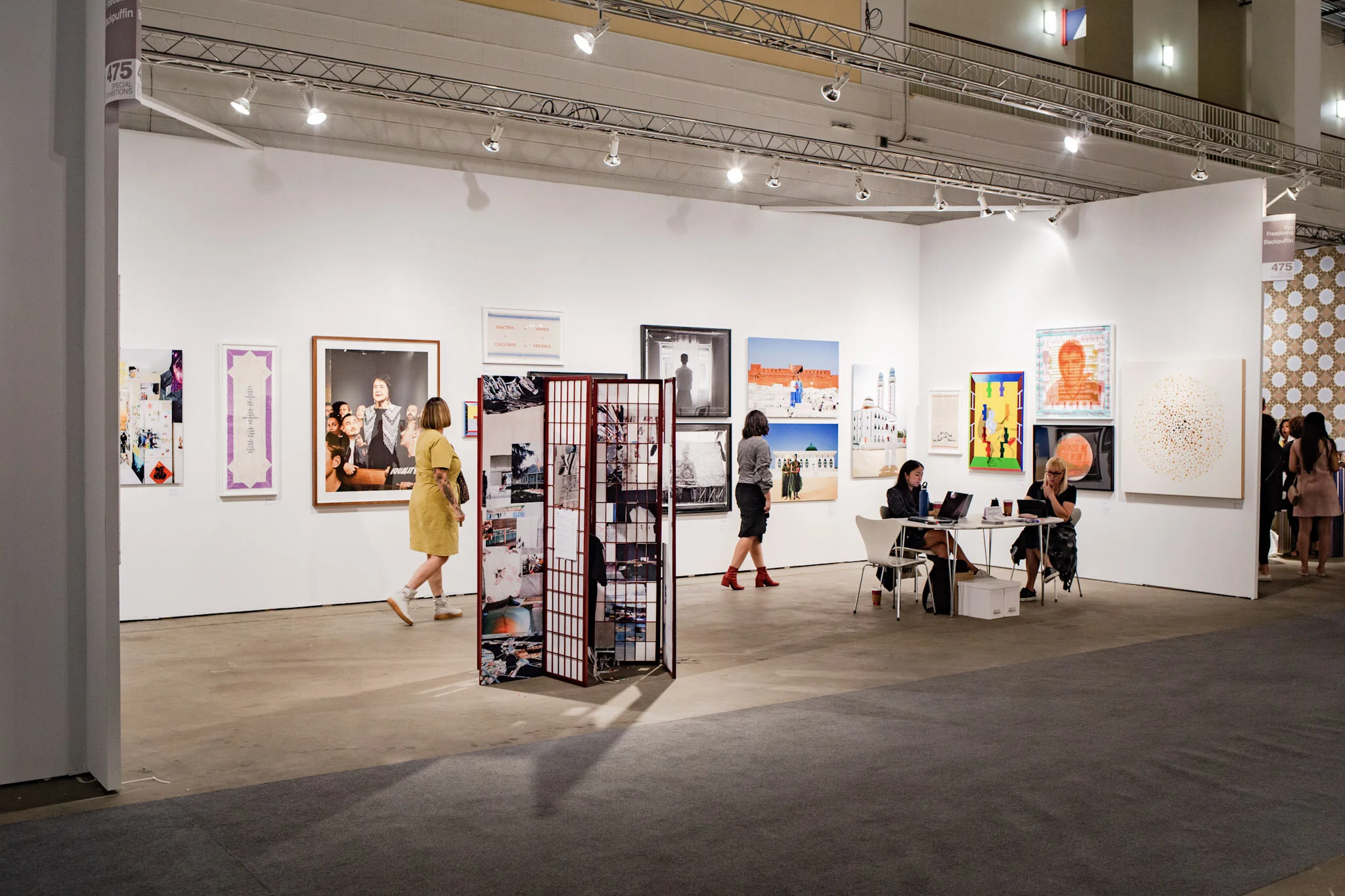 People browsing artwork at an art gallery or exhibition with various paintings and photographs displayed on white walls, and two women working at a table with laptops.