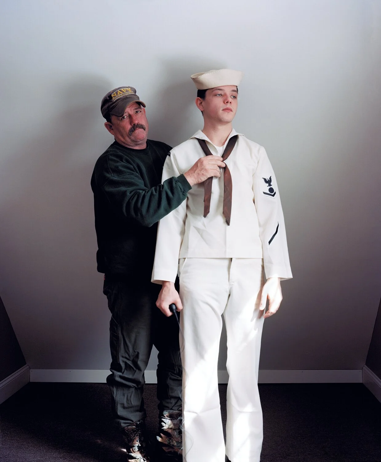 Man in navy uniform assistance adjusting a sailor's uniform with a white hat, white suit, and brown neckerchief.