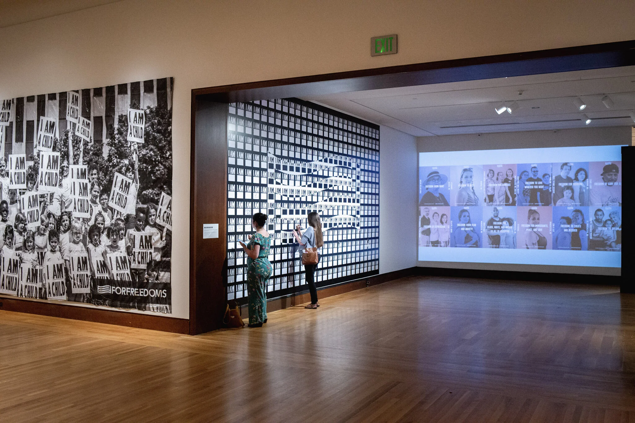 Two women exploring an art exhibit with large black-and-white photographs, including one with children holding signs that say "I AM A CHILD" and a blue-toned collage of portraits.