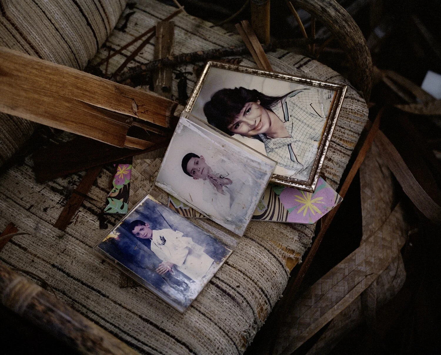Three framed photographs of young women placed on a worn, patterned fabric chair with wooden armrests, surrounded by torn newspaper and floral paper confetti.