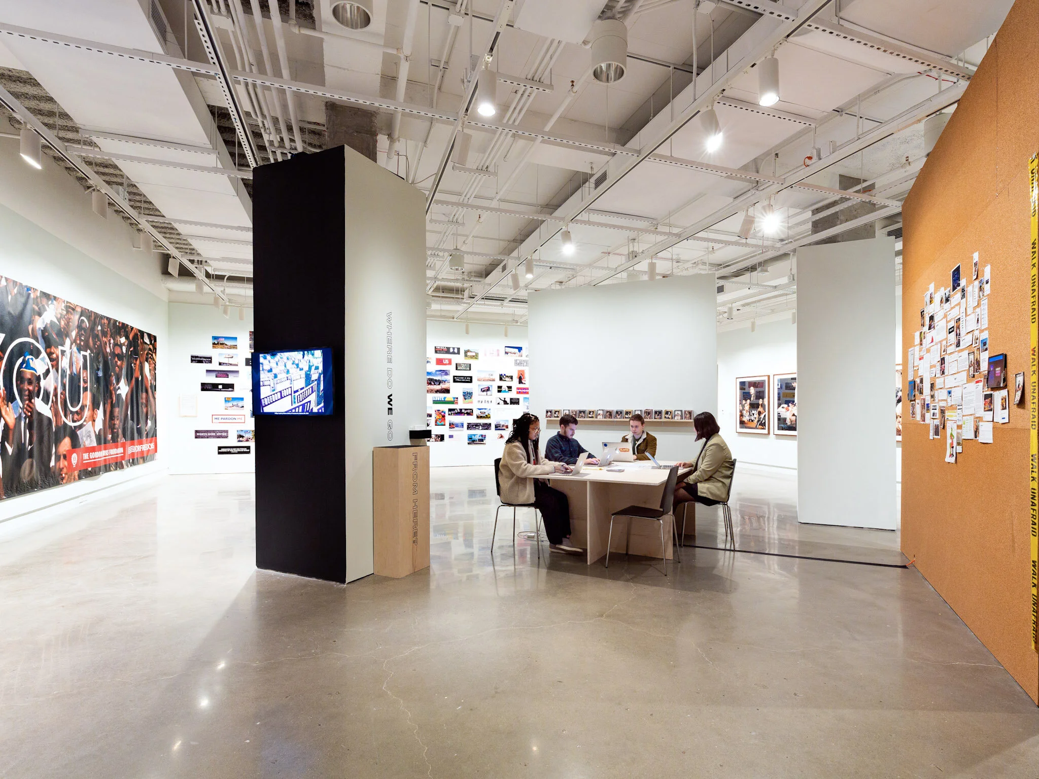 A group of four people sitting around a table in an art gallery or museum, looking at laptops and engaging in discussion. The gallery has white walls with various artworks, photographs, and installations displayed, and a polished concrete floor.