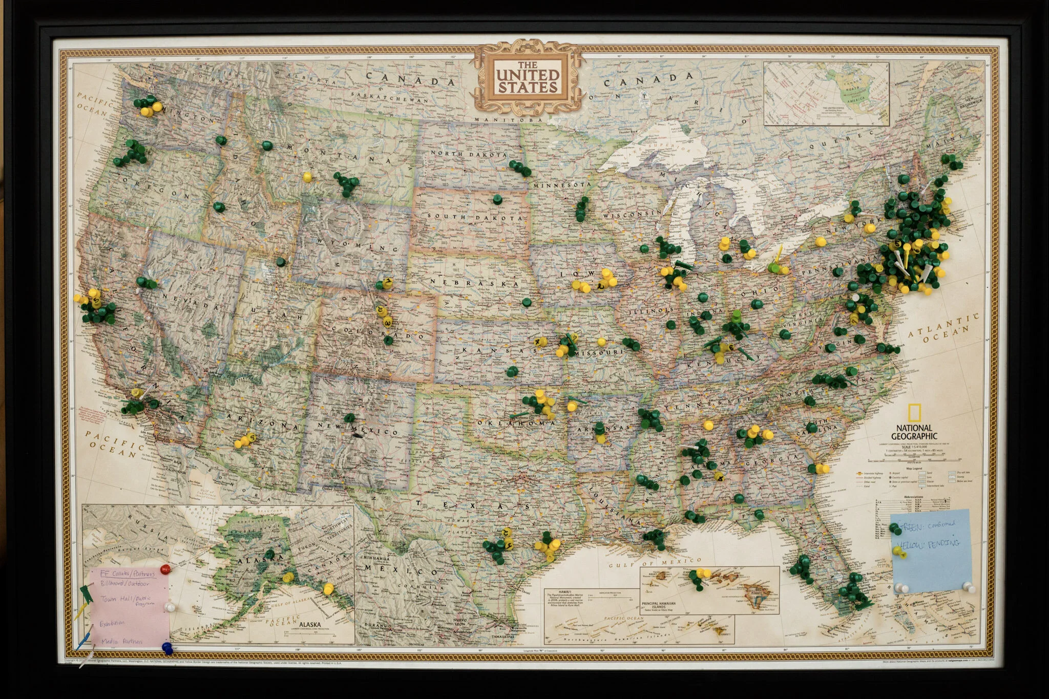 Framed map of the United States with various colored push pins marking locations. There are sticky notes on the bottom right and bottom left corners, and a few pins on Alaska.