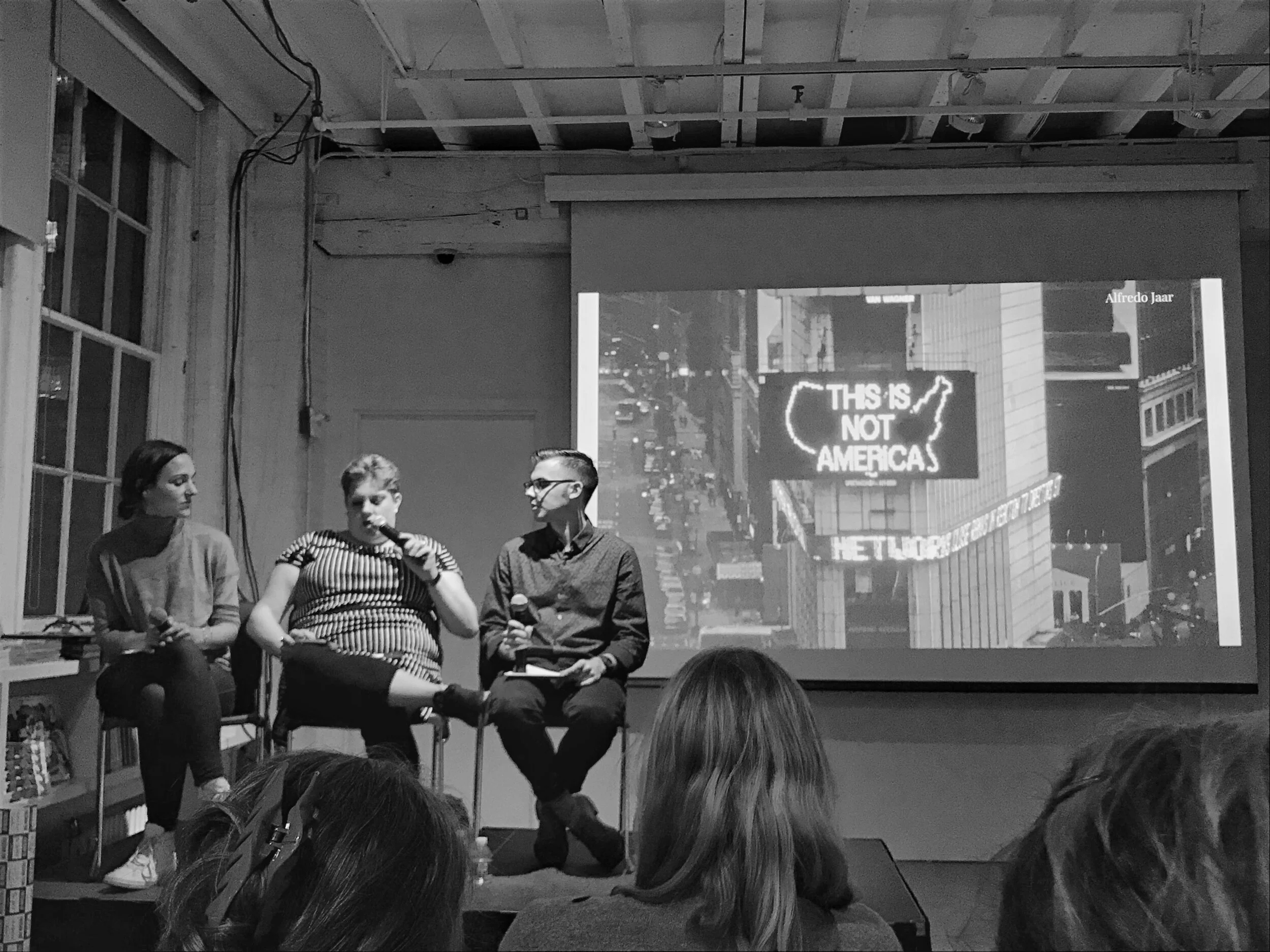 Three people sit on a stage in front of an audience, participating in a panel discussion. Behind them, a large screen displays an image of a cityscape with a neon sign that reads 'This is not America' and the name Alfredo Jaar.